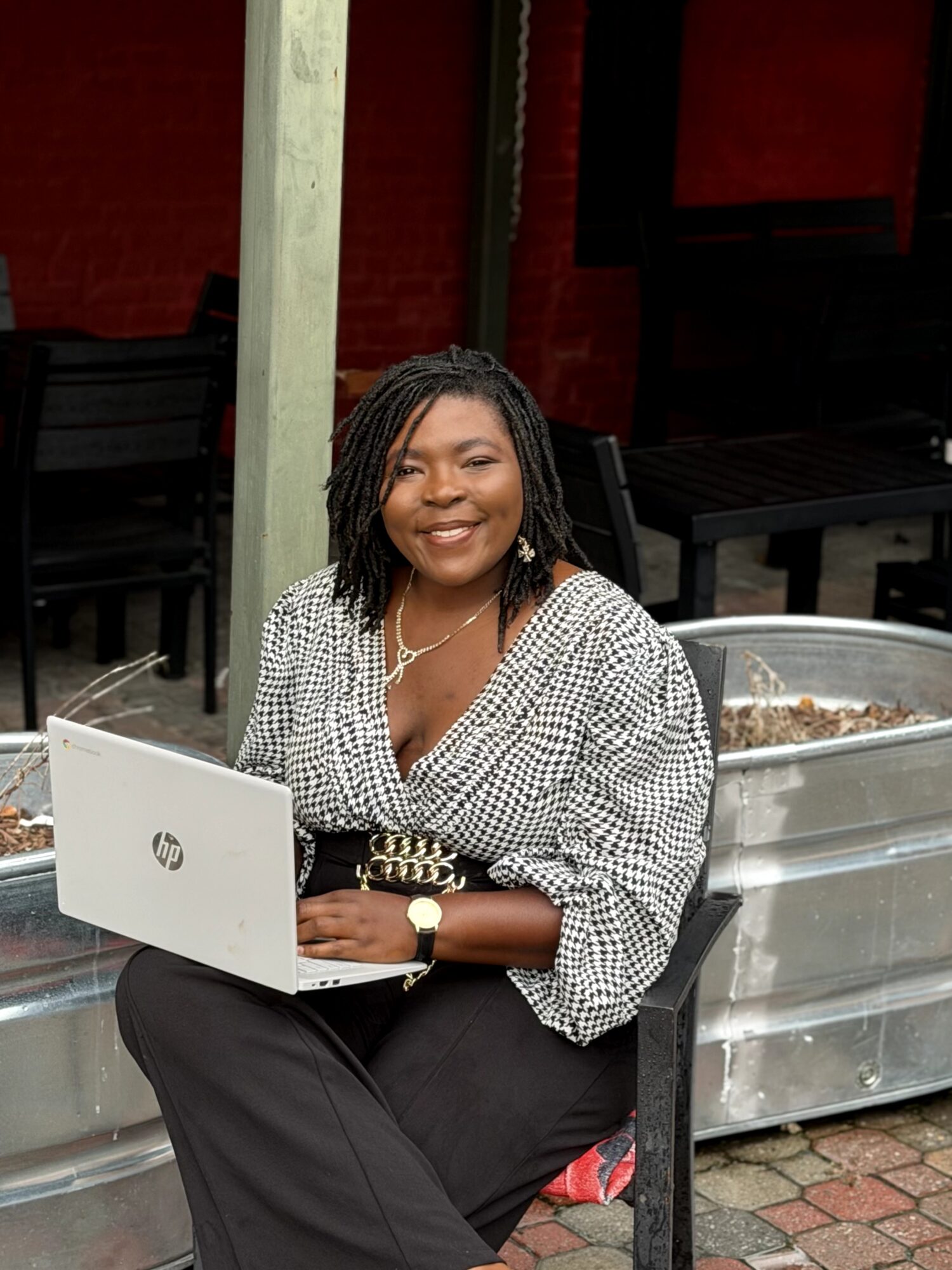 Smiling woman with dreadlocks sitting outdoors with a laptop, wearing a checkered blouse and black pants.