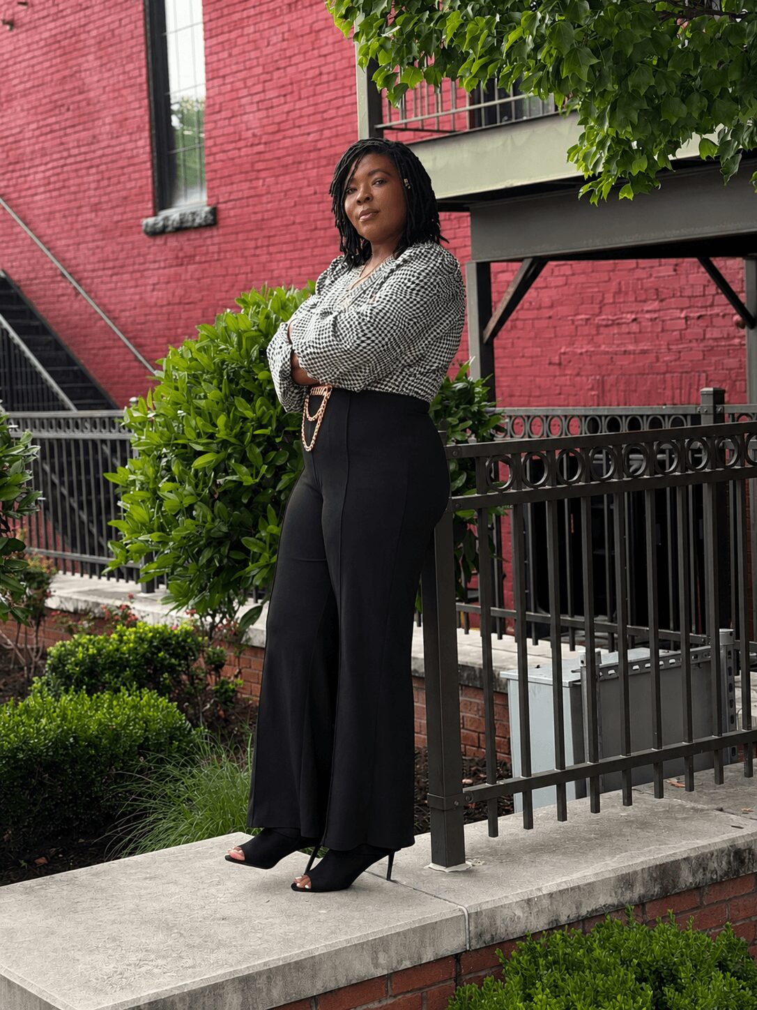 Woman standing outdoors near a black fence, with a red brick building and green trees in the background.