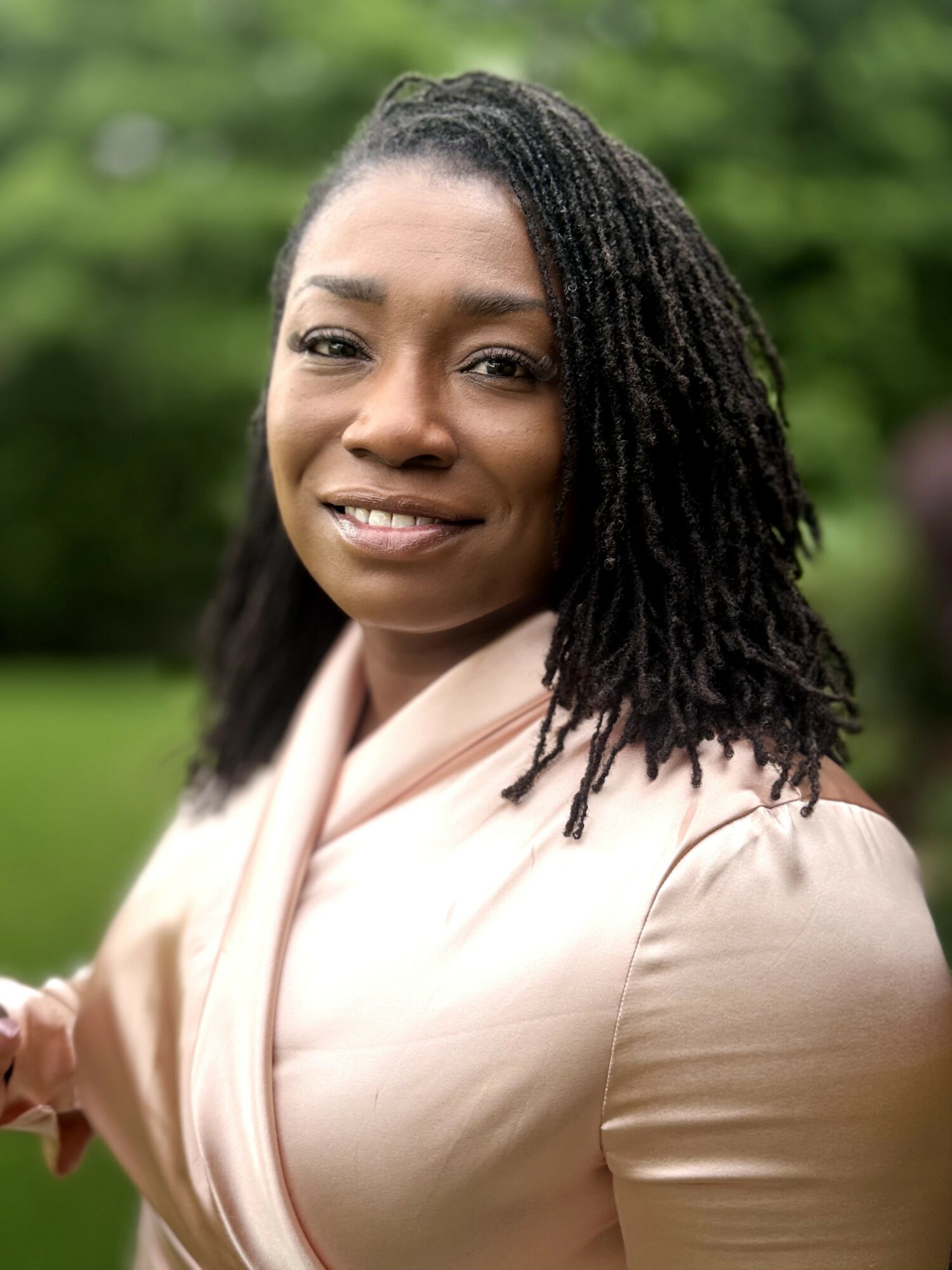 Portrait of a woman with shoulder-length dreadlocks, smiling outdoors with green trees in the background.
