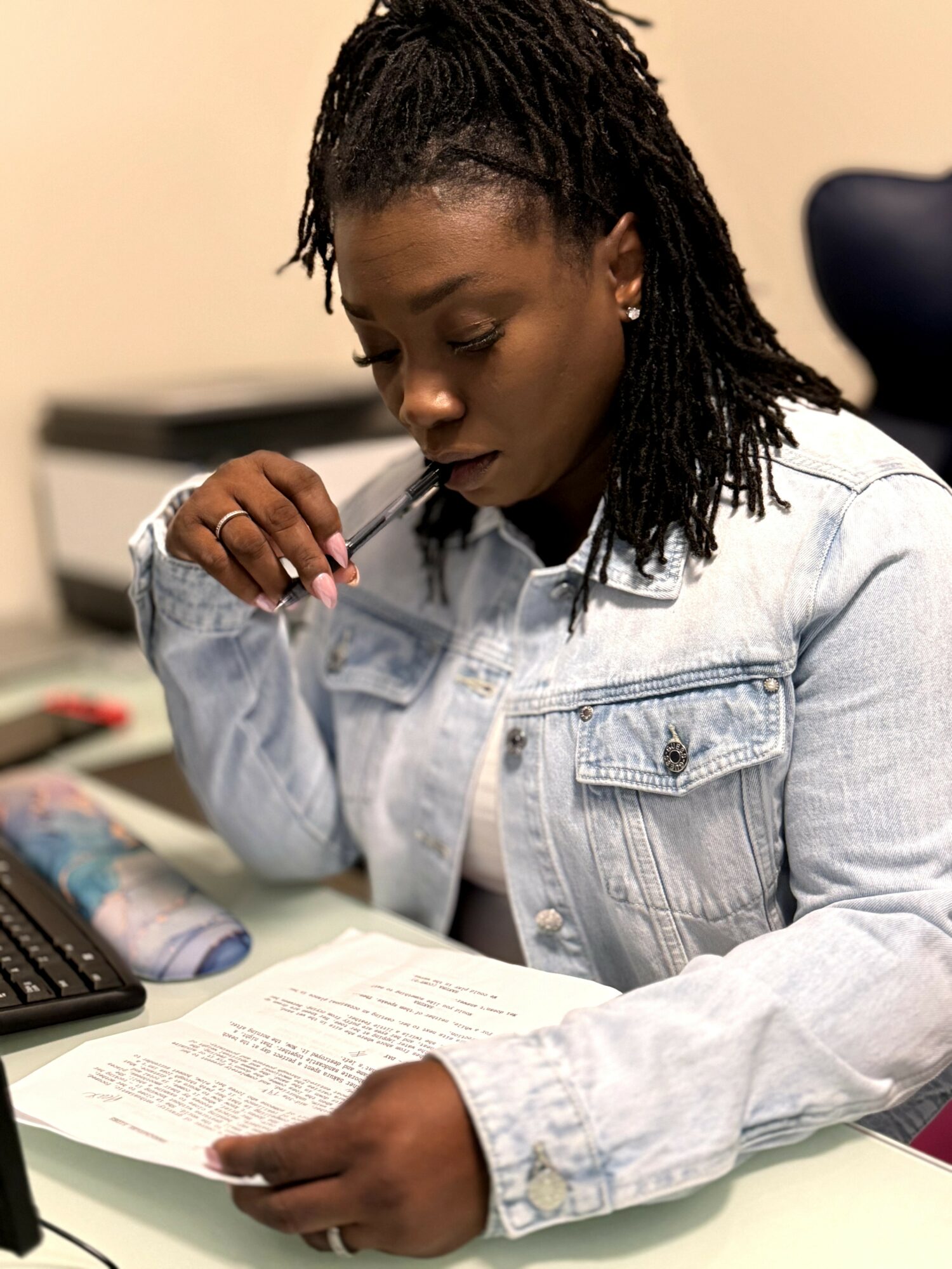 Woman reading a document at a desk, wearing a light denim jacket, with a computer and office supplies nearby.