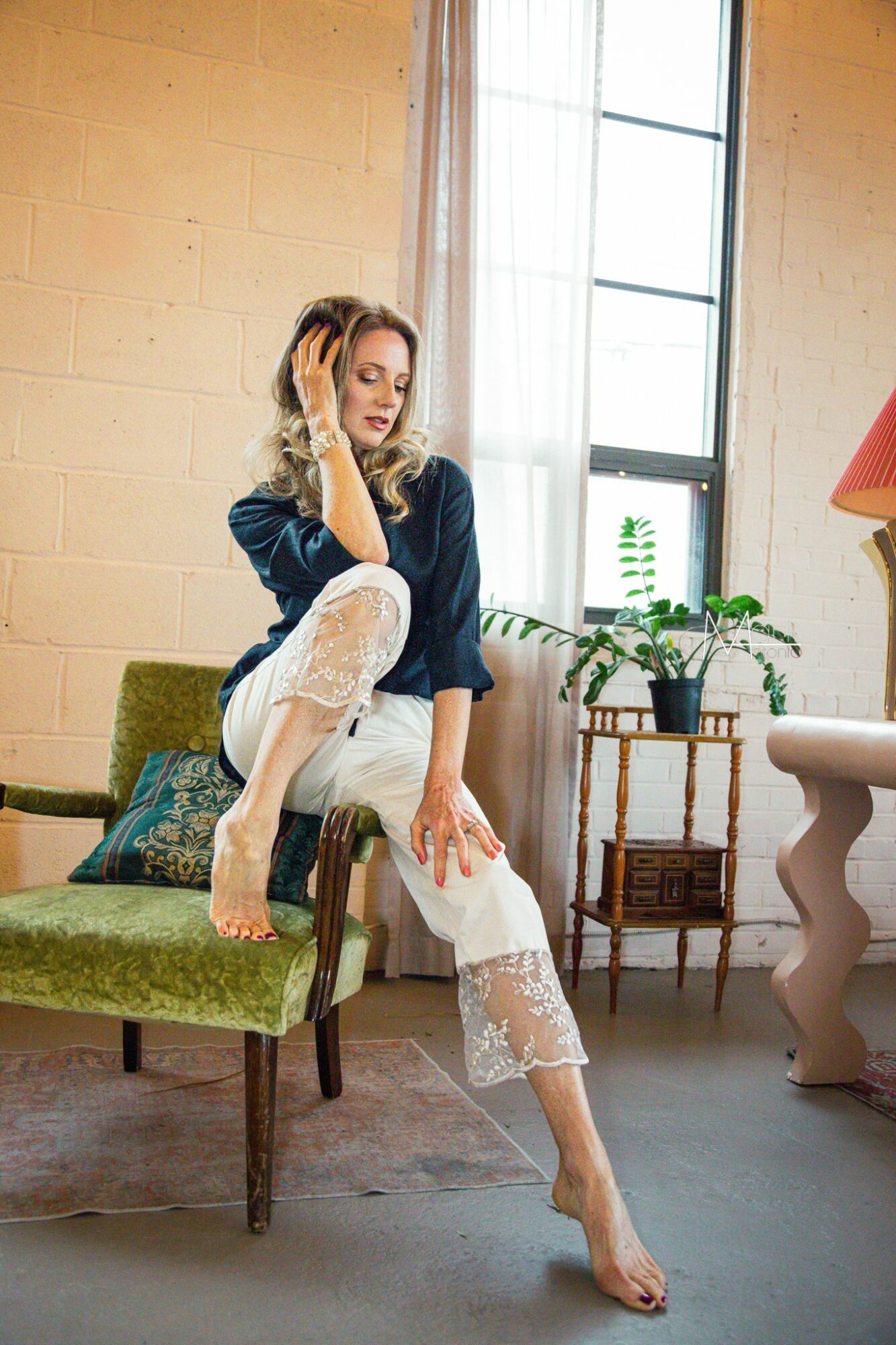Woman sitting on a green chair near a window, with plants and a lamp in the background, in a bright room.