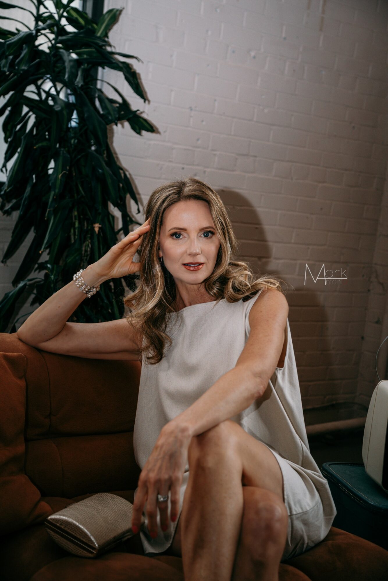 Woman with wavy hair sitting on a brown sofa, wearing a white sleeveless dress, with a large plant and white brick wall behind her.