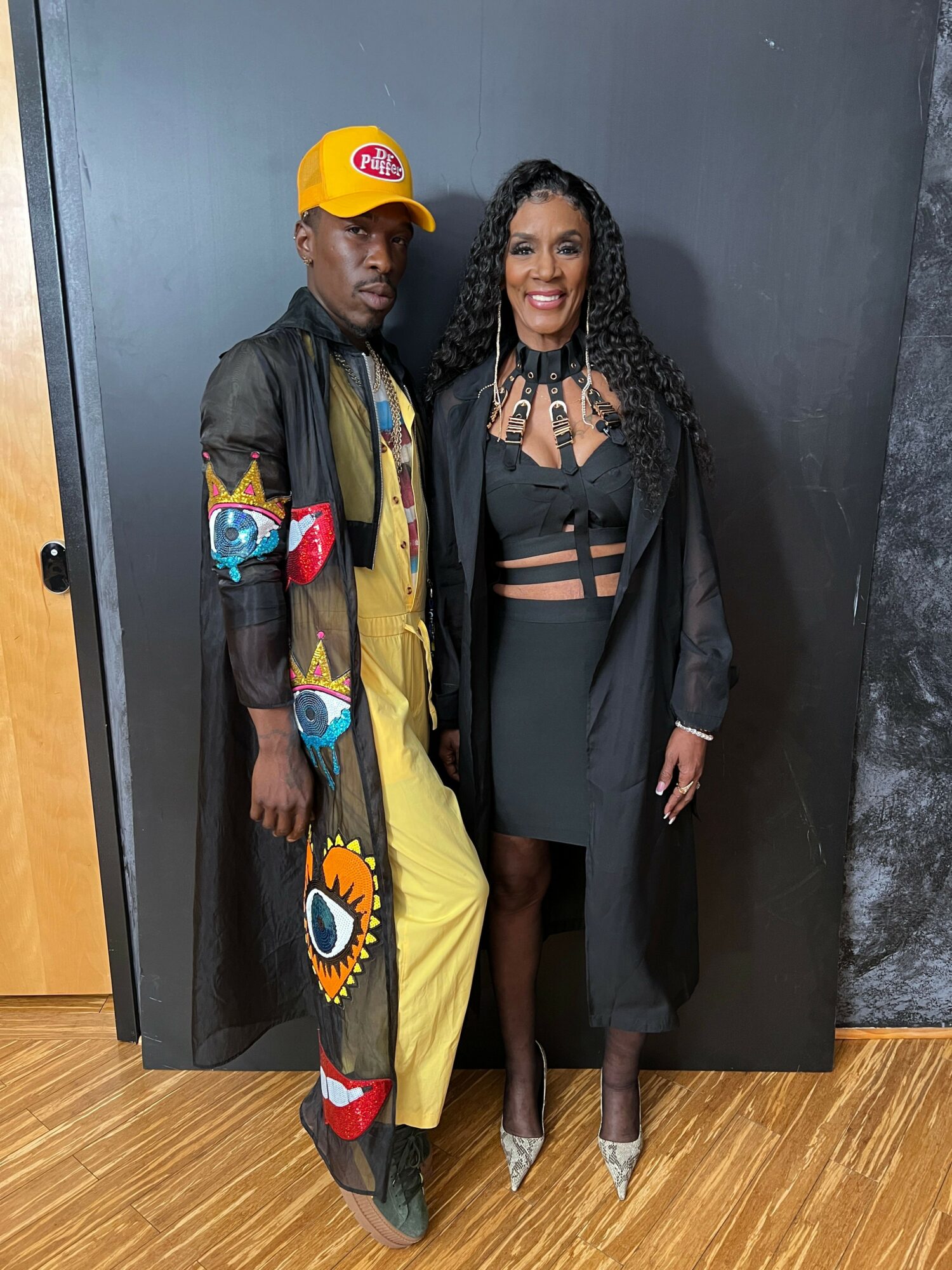 Two women standing against a black backdrop, one in a black outfit with a jacket, the other in a colorful jacket and yellow cap, on a wooden floor.
