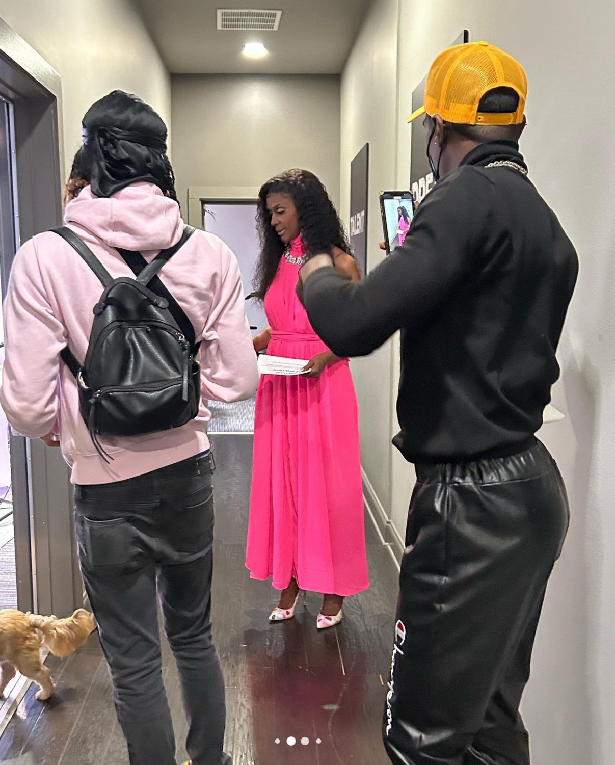 Three people in a hallway, one woman in a pink dress, two men with backpacks, and a small dog nearby.