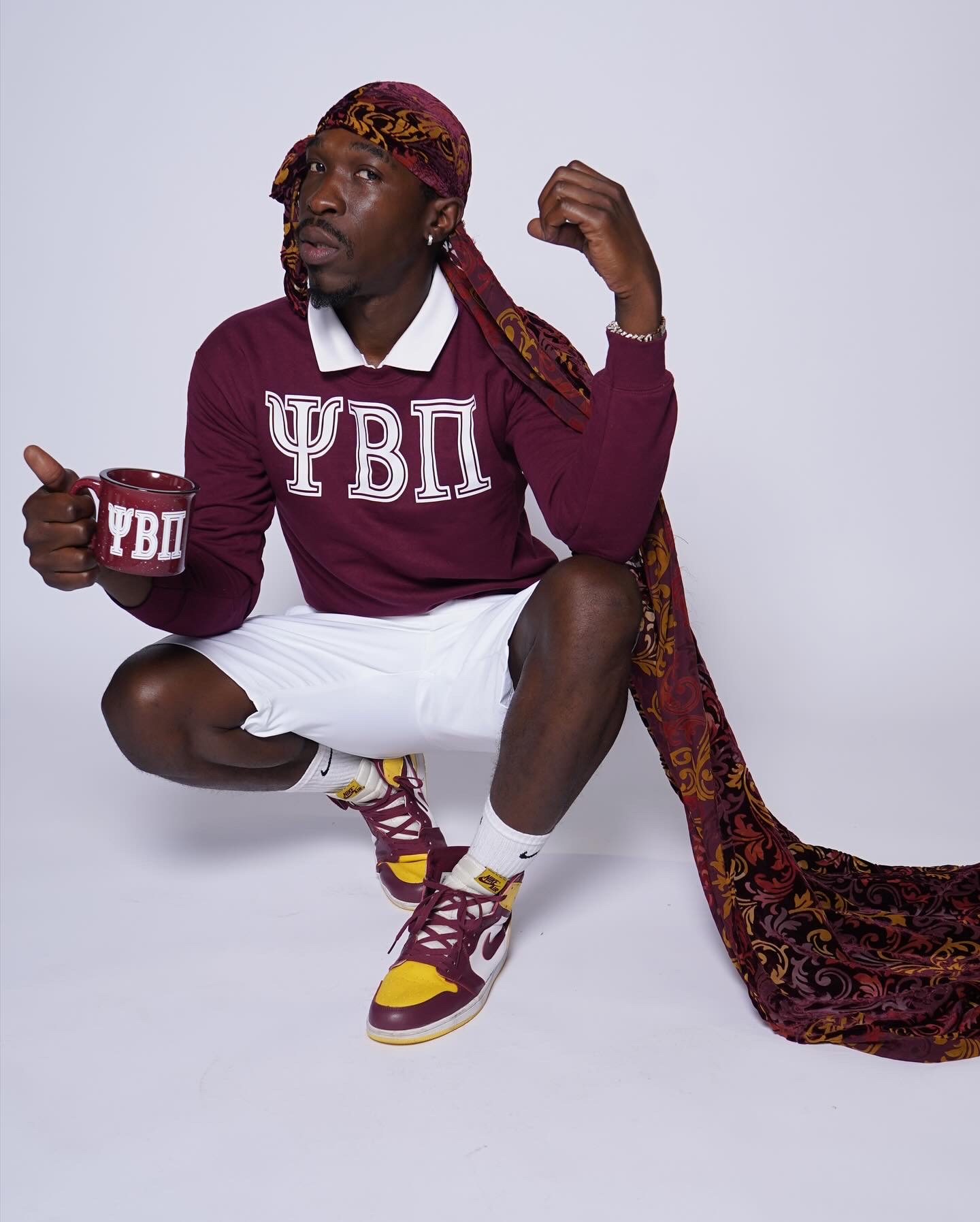 Person crouching, wearing a maroon sweatshirt with Greek letters, holding a mug, and wearing a patterned headscarf and sneakers.
