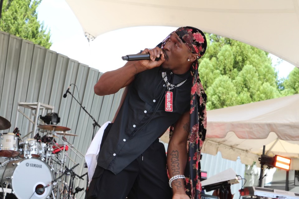 Person singing into a microphone outdoors under a canopy, with musical instruments in the background.