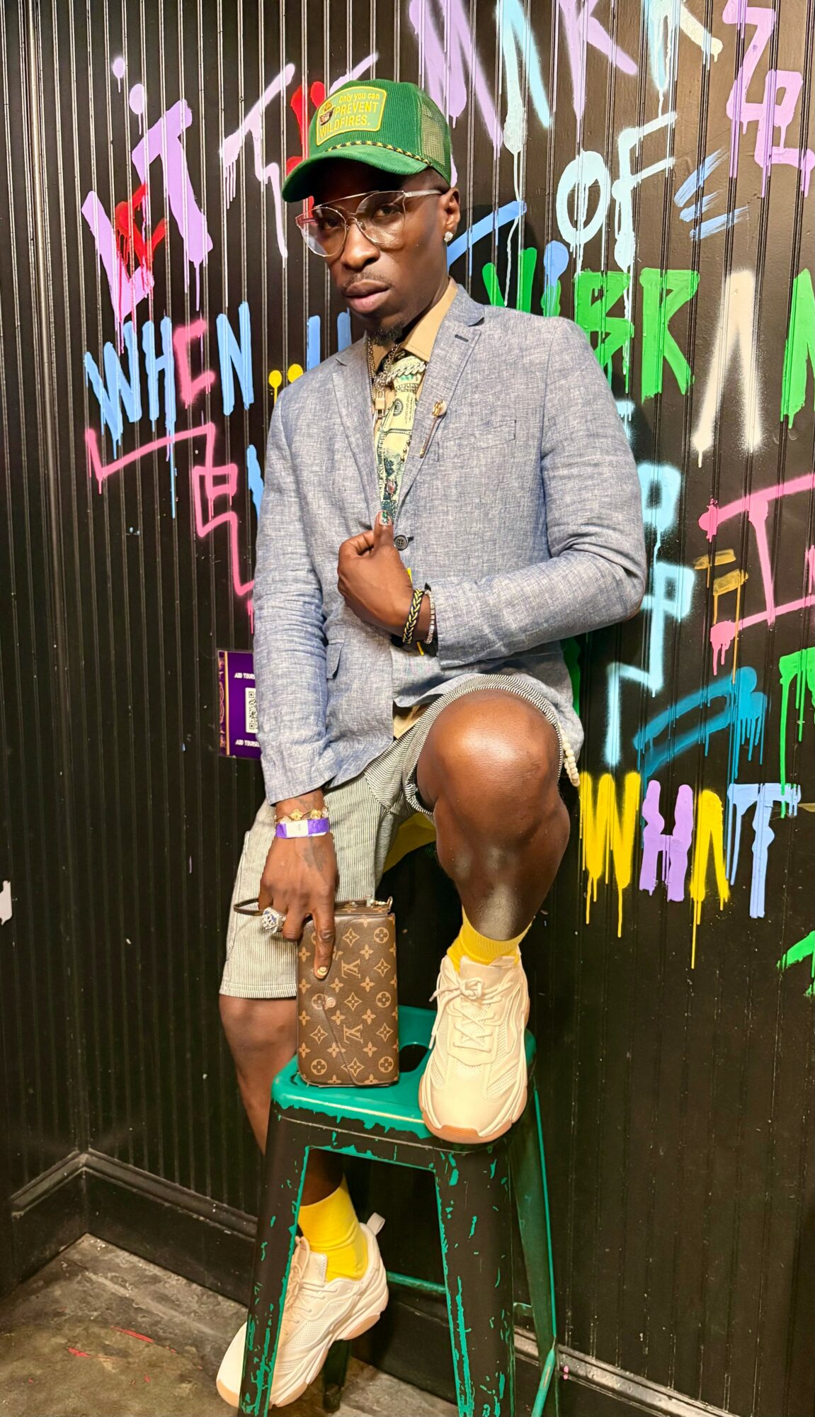 Person wearing a gray blazer, shorts, yellow shoes, and a green cap, standing against a colorful graffiti wall.