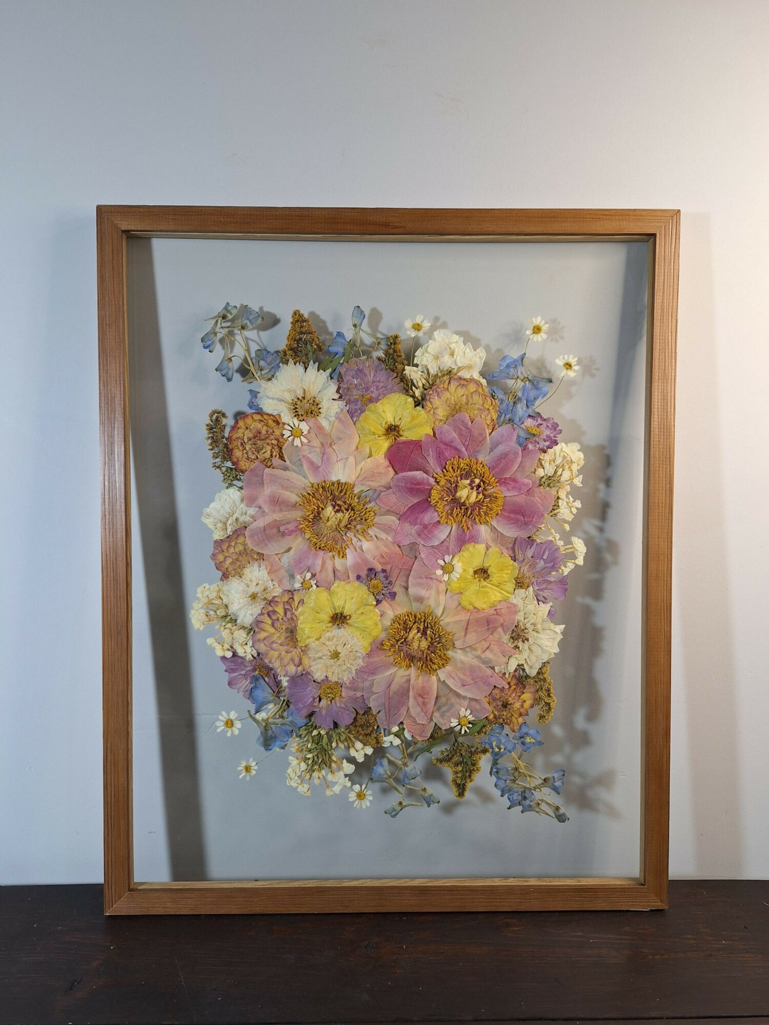Framed floral artwork with pink, yellow, and white flowers, viewed at an angle, with a wooden frame and glass cover.