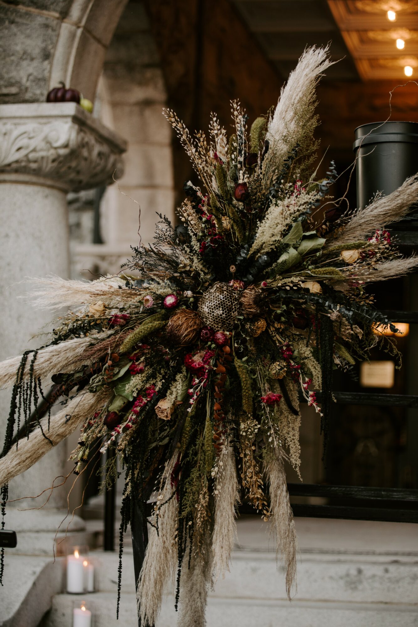 A large floral arrangement with dried and fresh flowers, grasses, and berries, hanging indoors with candles nearby.