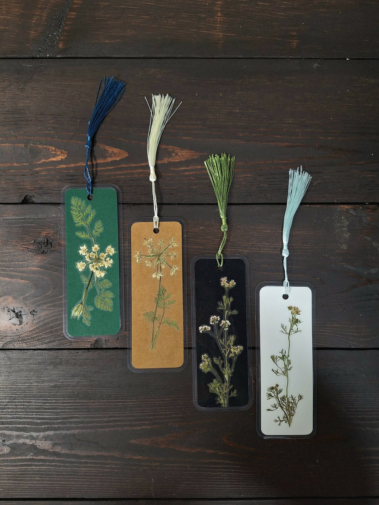 Four botanical tags with tassels, each featuring different plant illustrations, arranged on a dark wooden surface.