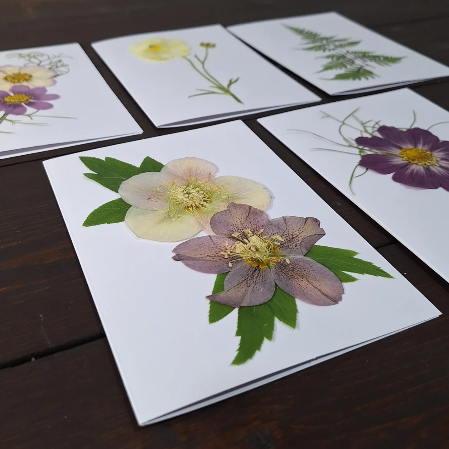 Pressed flowers arranged on white paper, with green leaves, on a dark wooden surface.