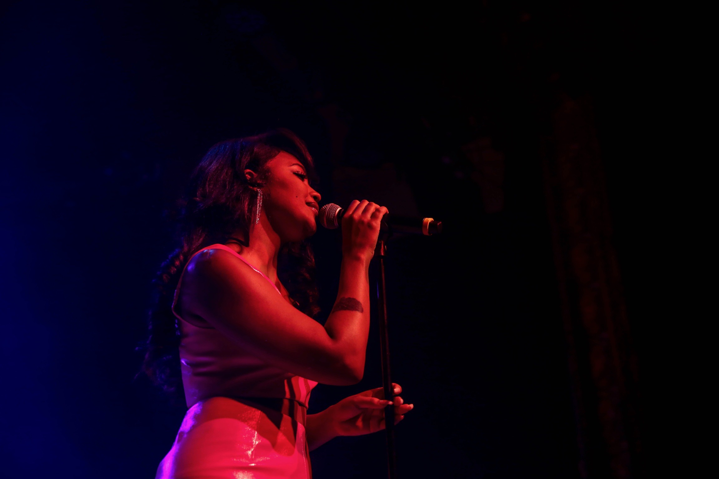 Woman singing into a microphone on stage with dark background, red and blue lighting, holding a glass, long hair, sleeveless top