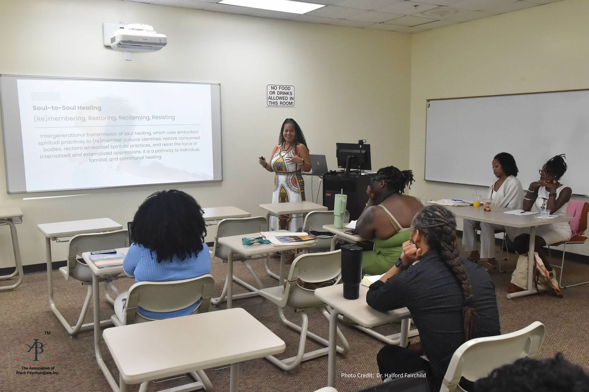 A woman stands at the front of a classroom giving a presentation to seated students, with a whiteboard and projector screen behind her.