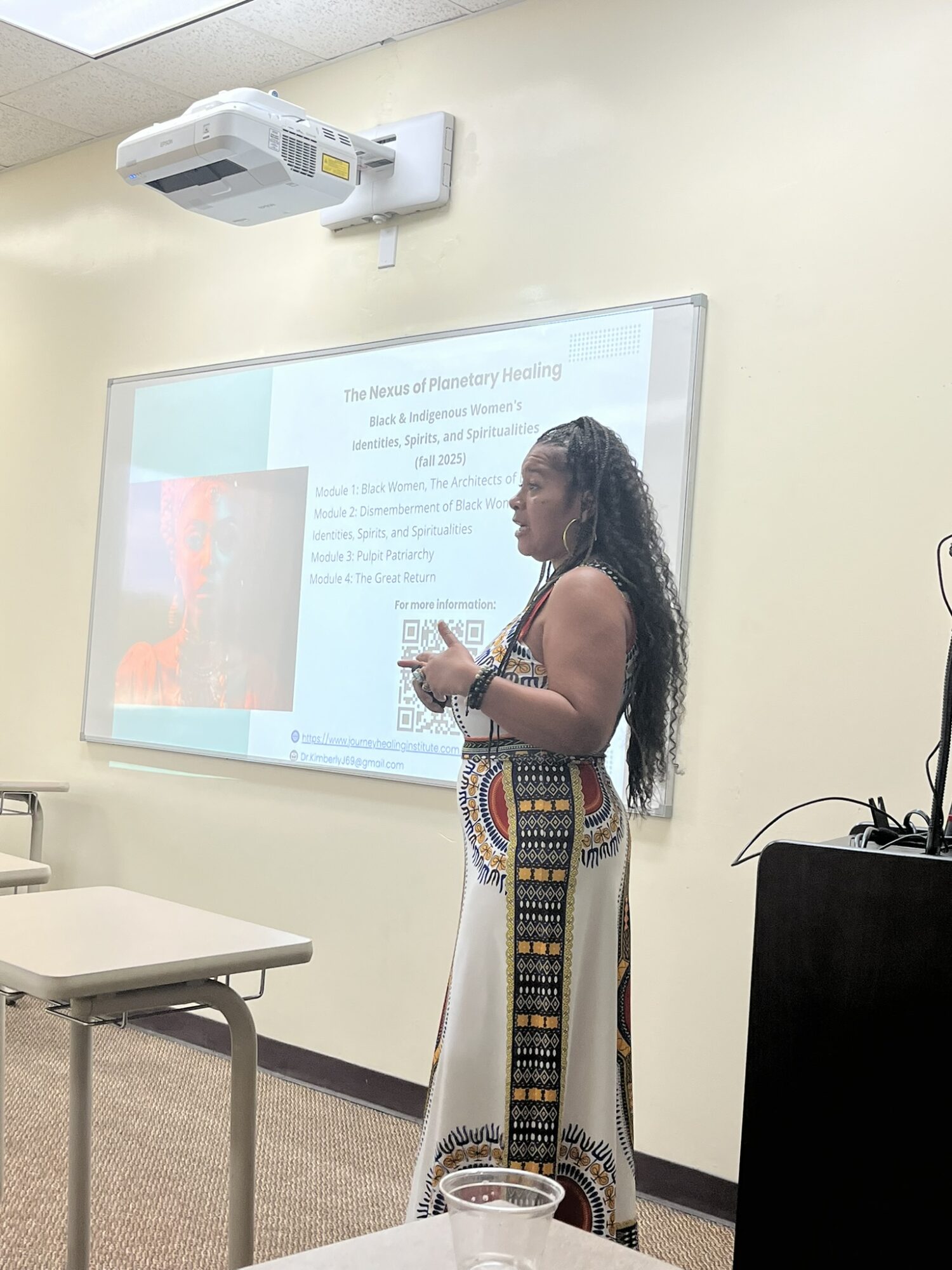 Woman standing in front of a whiteboard with a presentation slide, holding a marker, in a classroom.