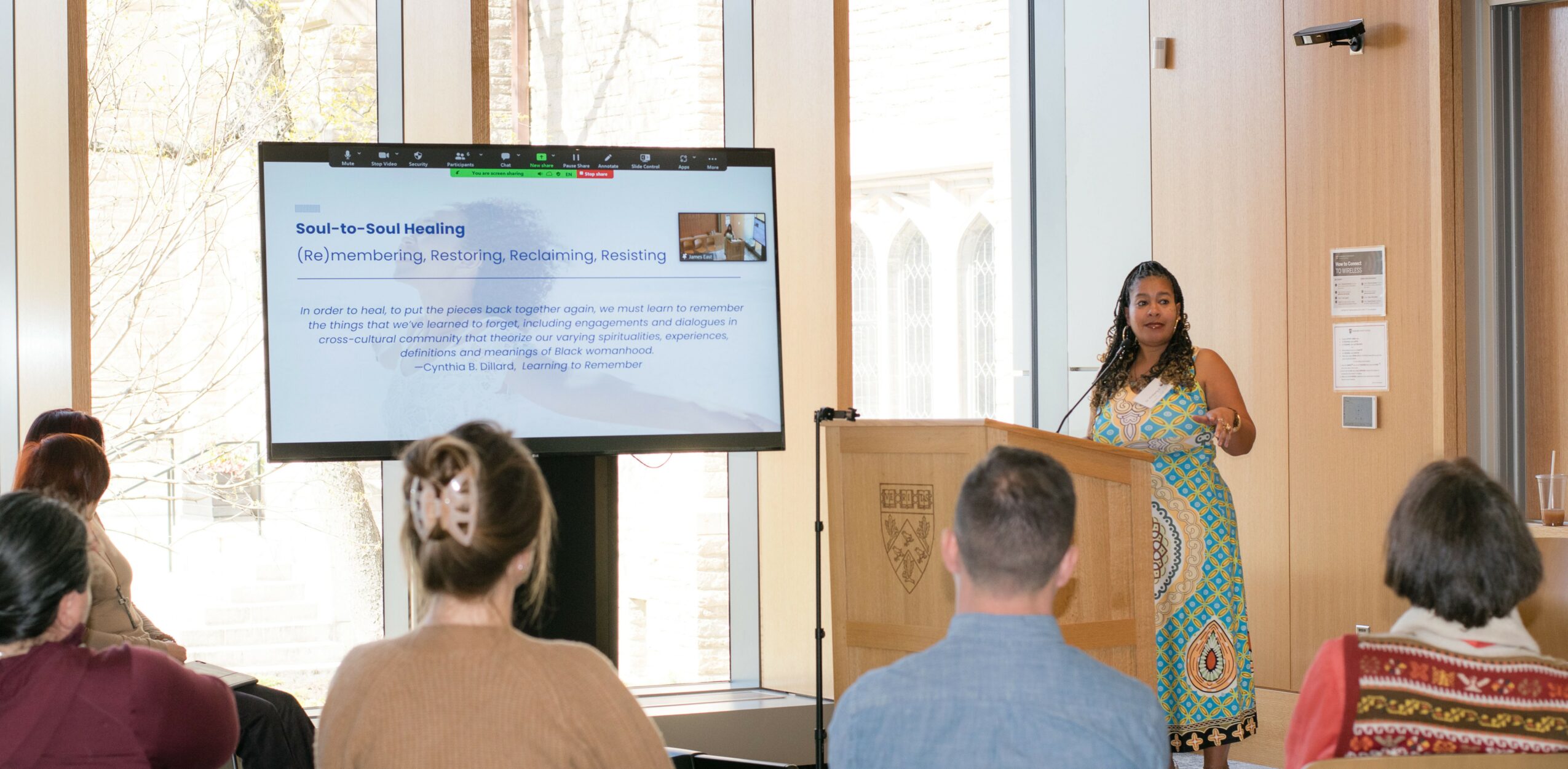Woman presenting in front of a large screen to a seated audience in a bright room.