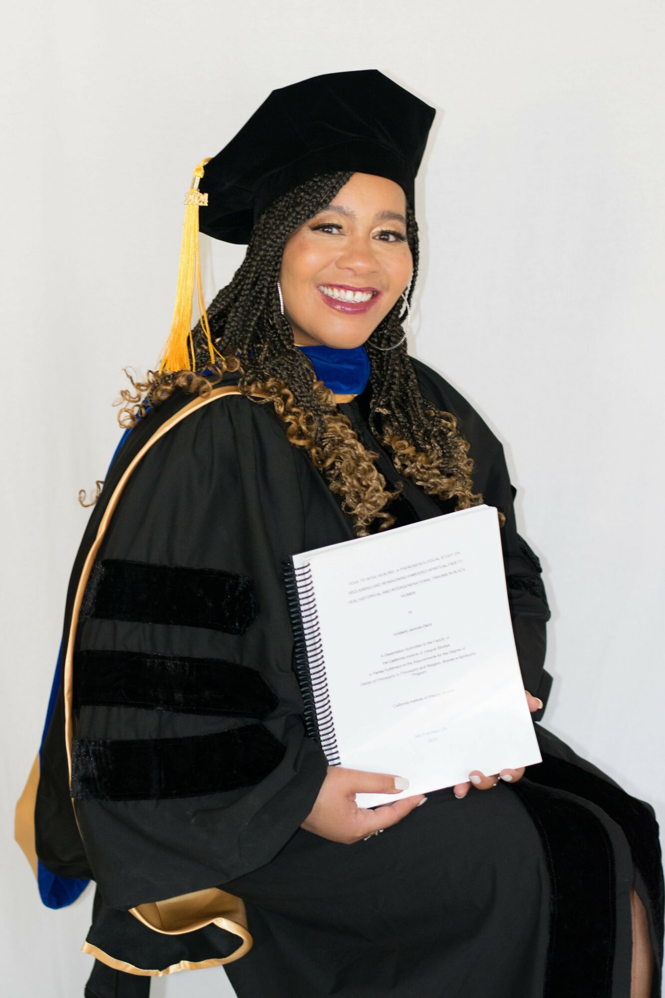 Woman in graduation gown and cap holding a diploma, smiling, against a plain background.