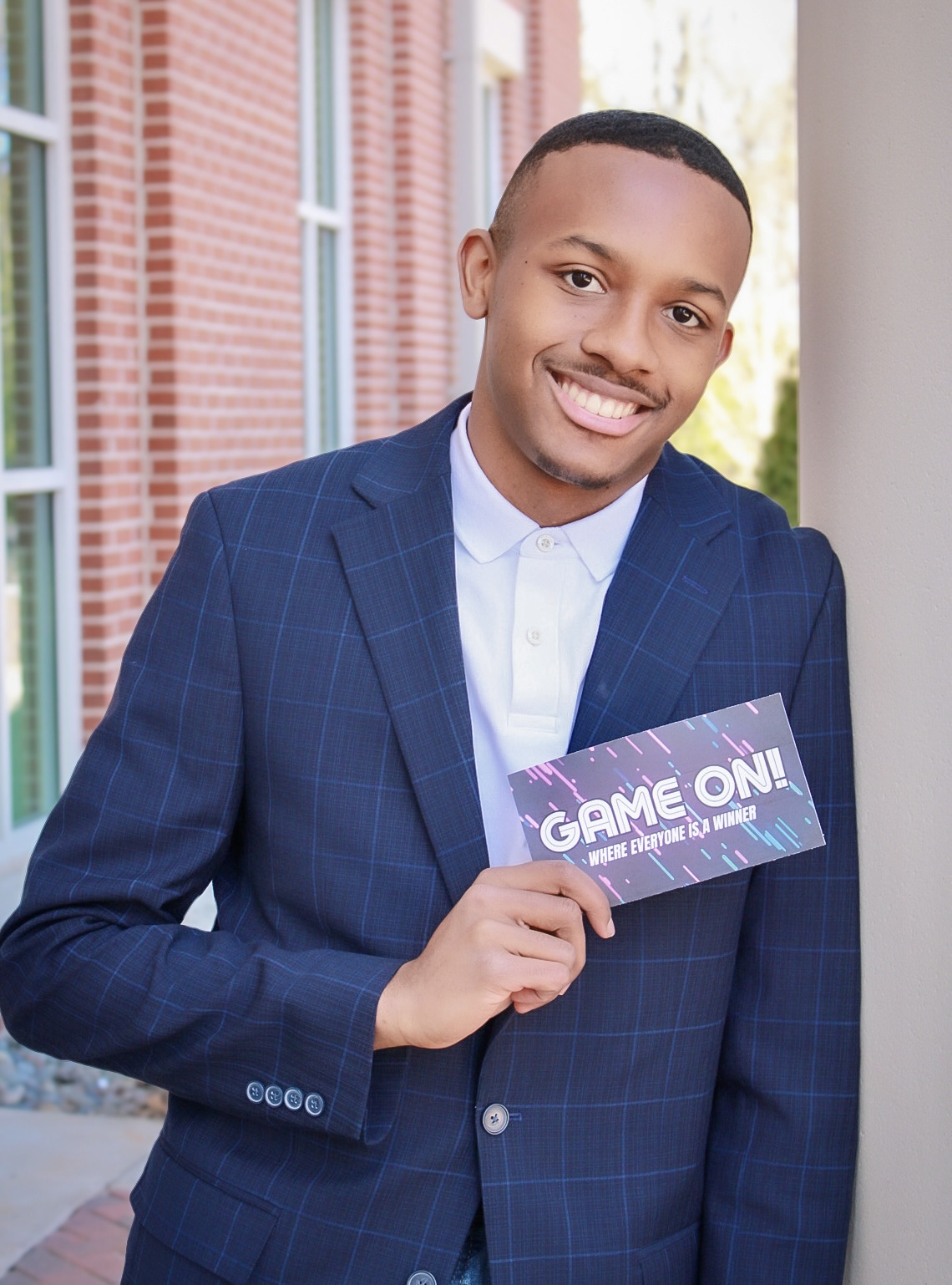 Young man in a suit holding a card that says 'GAME ON!' outdoors, smiling, brick building in background.