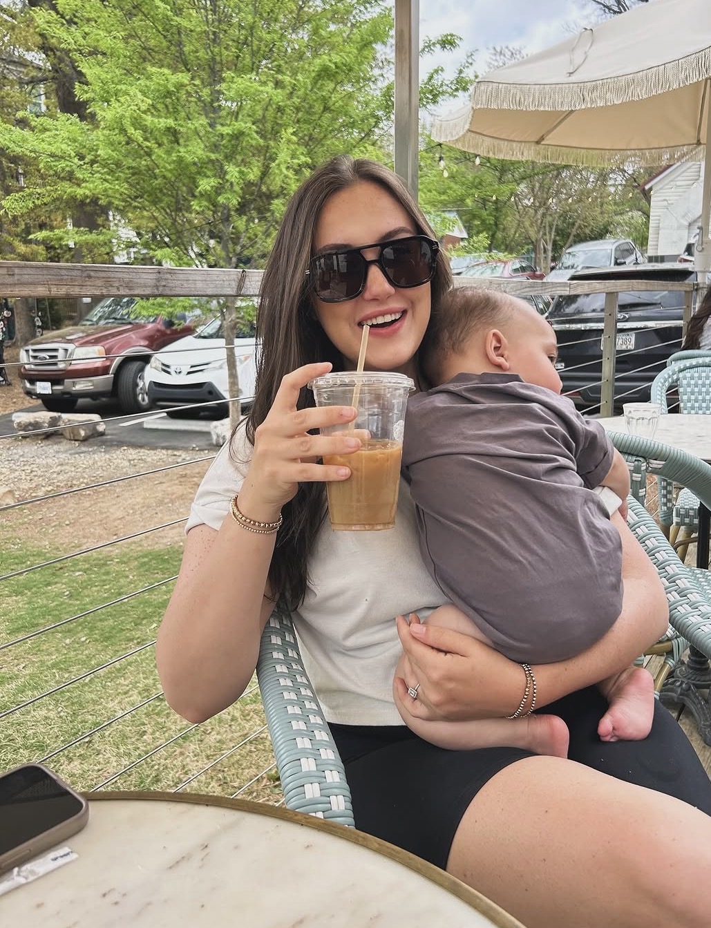 Woman with sunglasses smiling, holding a drink, sitting outdoors with a child in a gray shirt on her lap, trees and parked cars in background.