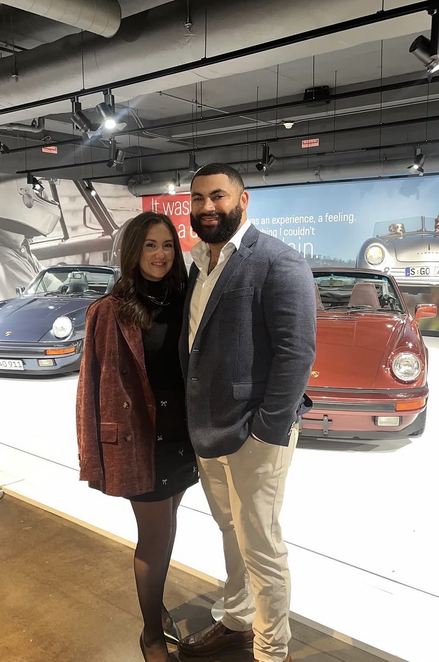A man and woman stand together in front of vintage cars inside a museum, smiling at the camera.