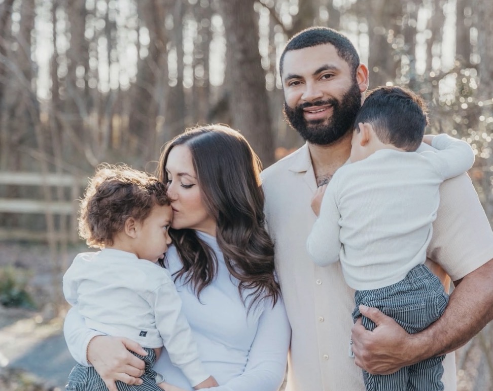 Family of four outdoors, woman holding a child, man holding another child, trees in background, sunlight filtering through.