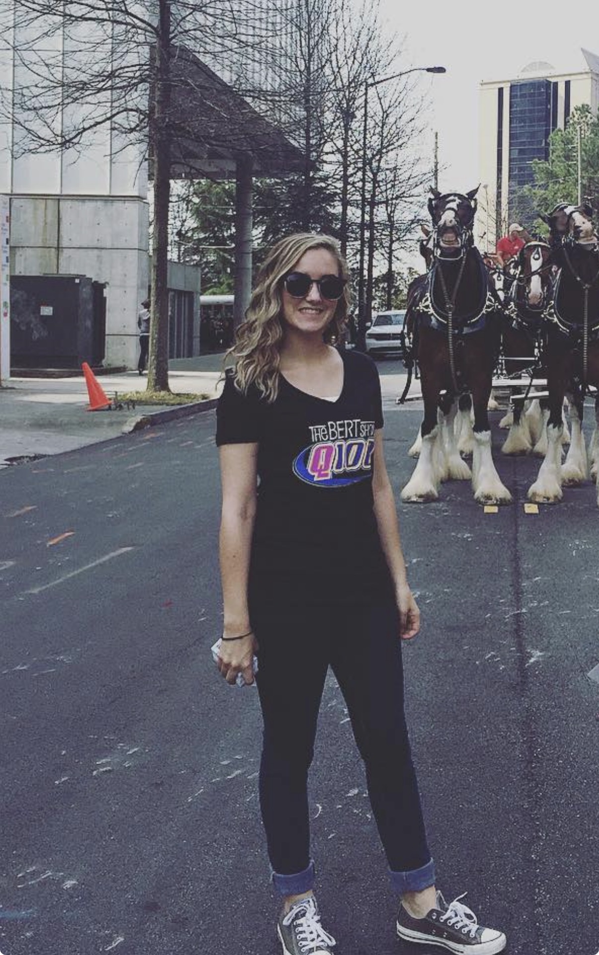 Young woman with sunglasses and wavy hair standing on street, horse-drawn carriages in background, urban setting.