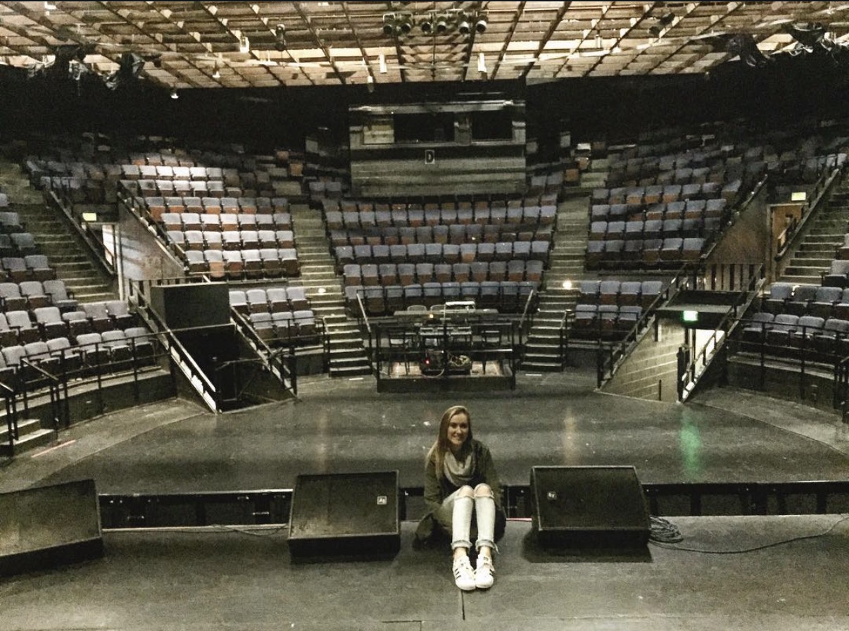 Person sitting on stage in an empty theater with multiple seating sections and stairs, facing forward.