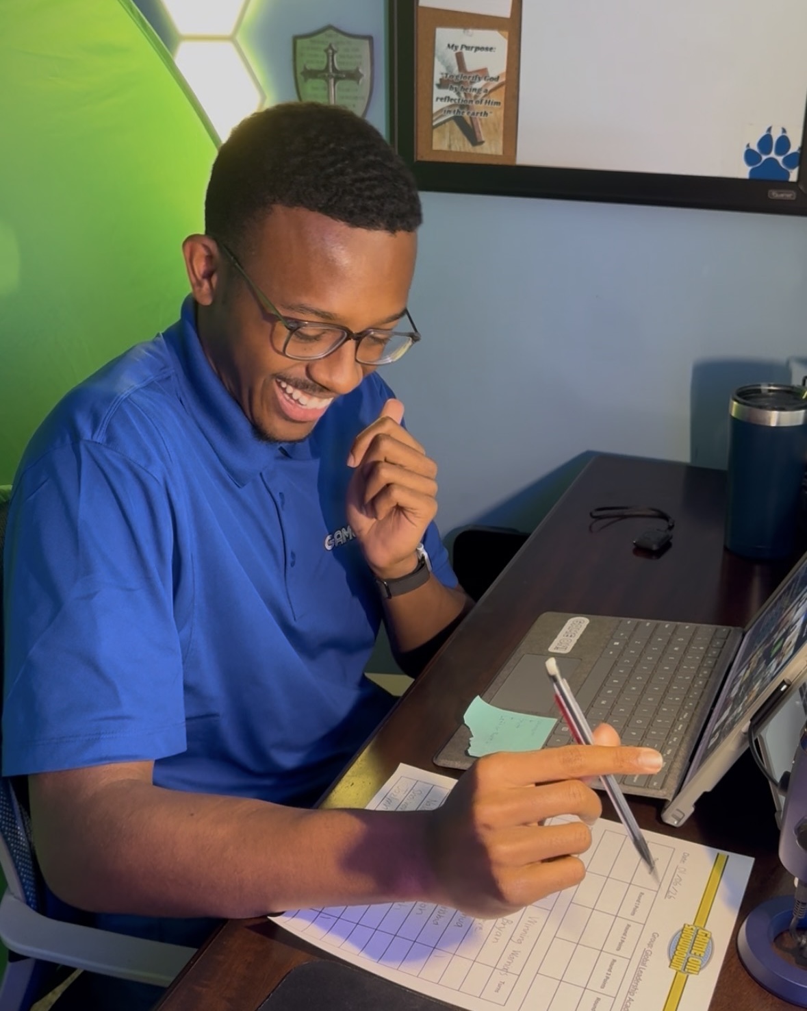 Young man in blue shirt smiling, sitting at a desk with a laptop, pointing at a tablet, in a bright room.