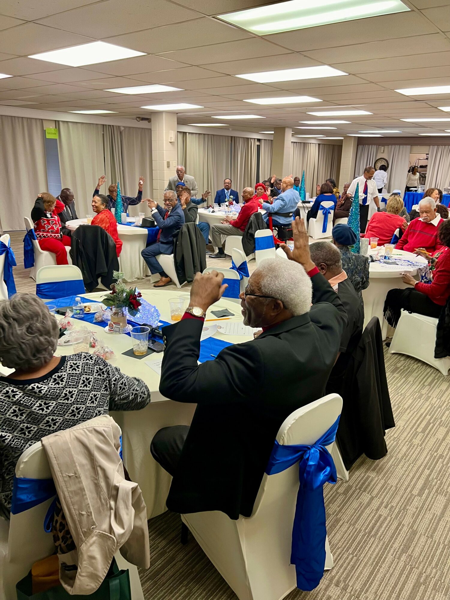People seated at round tables in a banquet hall, some raising glasses, with white tablecloths and blue chair sashes.