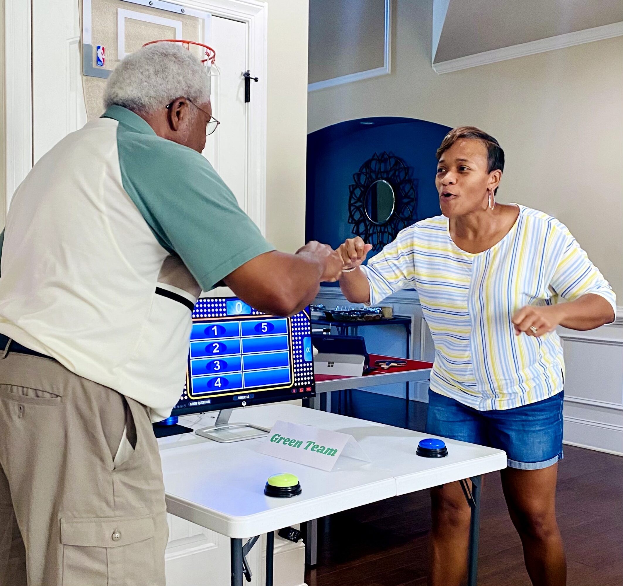 An elderly man and a woman shake hands in a game show setting with a screen and buzzer on the table.