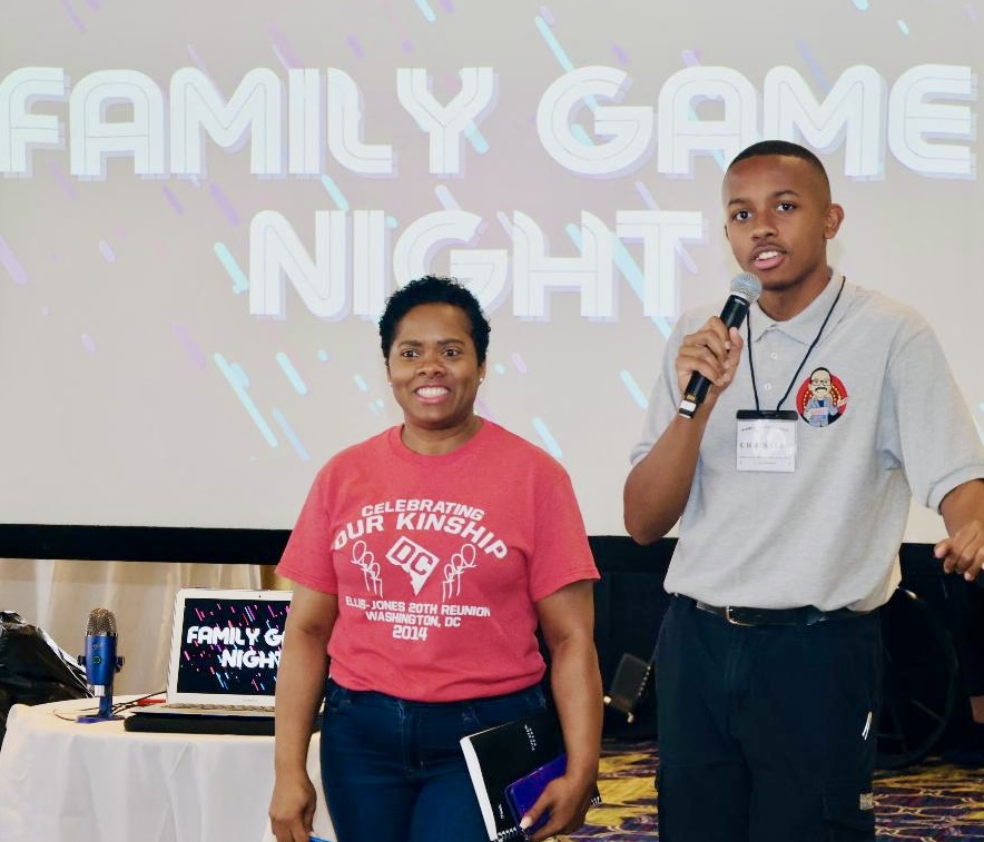 Two people standing in front of a screen at a family game night event, one holding a microphone, smiling.