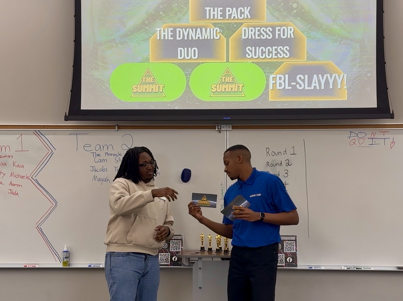 Two students stand in front of a whiteboard and screen, exchanging a small object and a book in a classroom.
