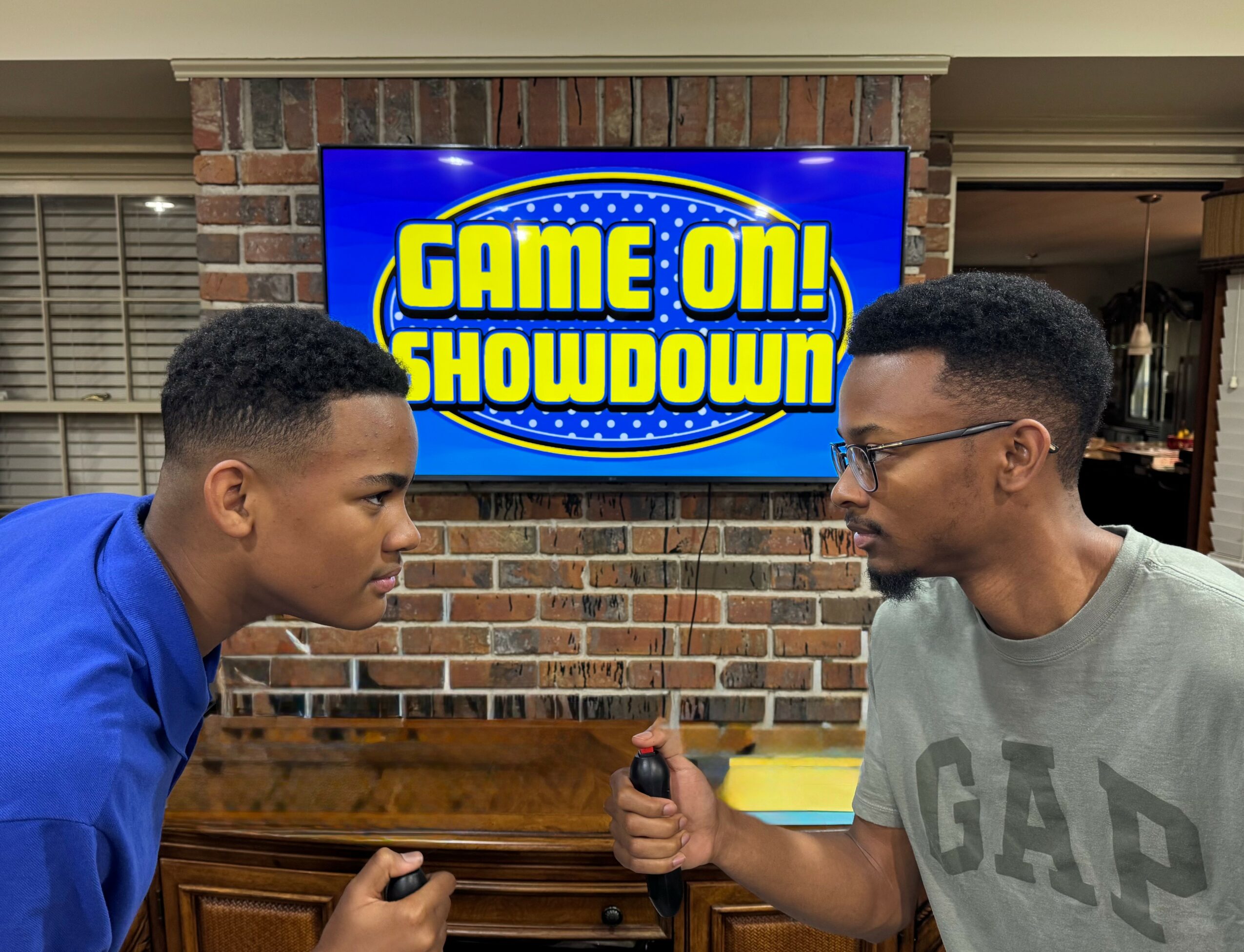 Two young men face each other with game controllers, preparing for a game showdown, in front of a screen that says 'Game On! Showdown'.