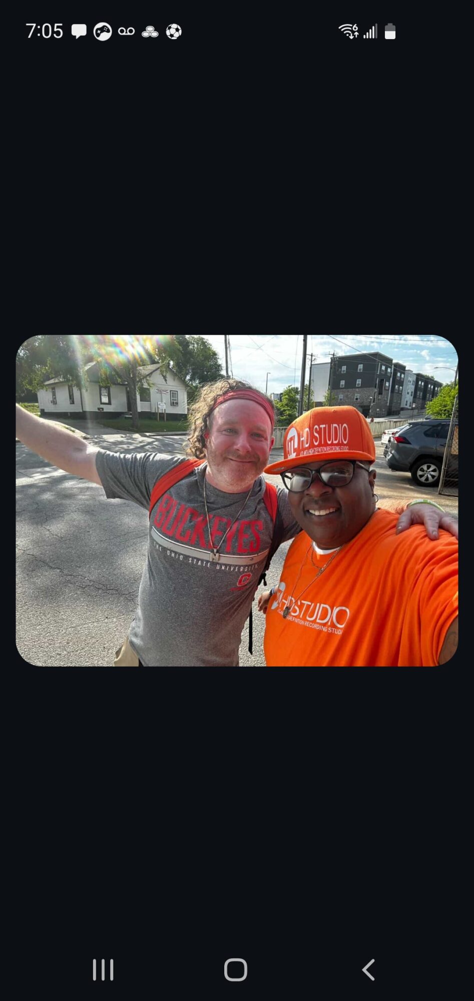 Two people smiling outdoors, one wearing a gray shirt and the other in an orange shirt and hat, with trees and buildings in background.