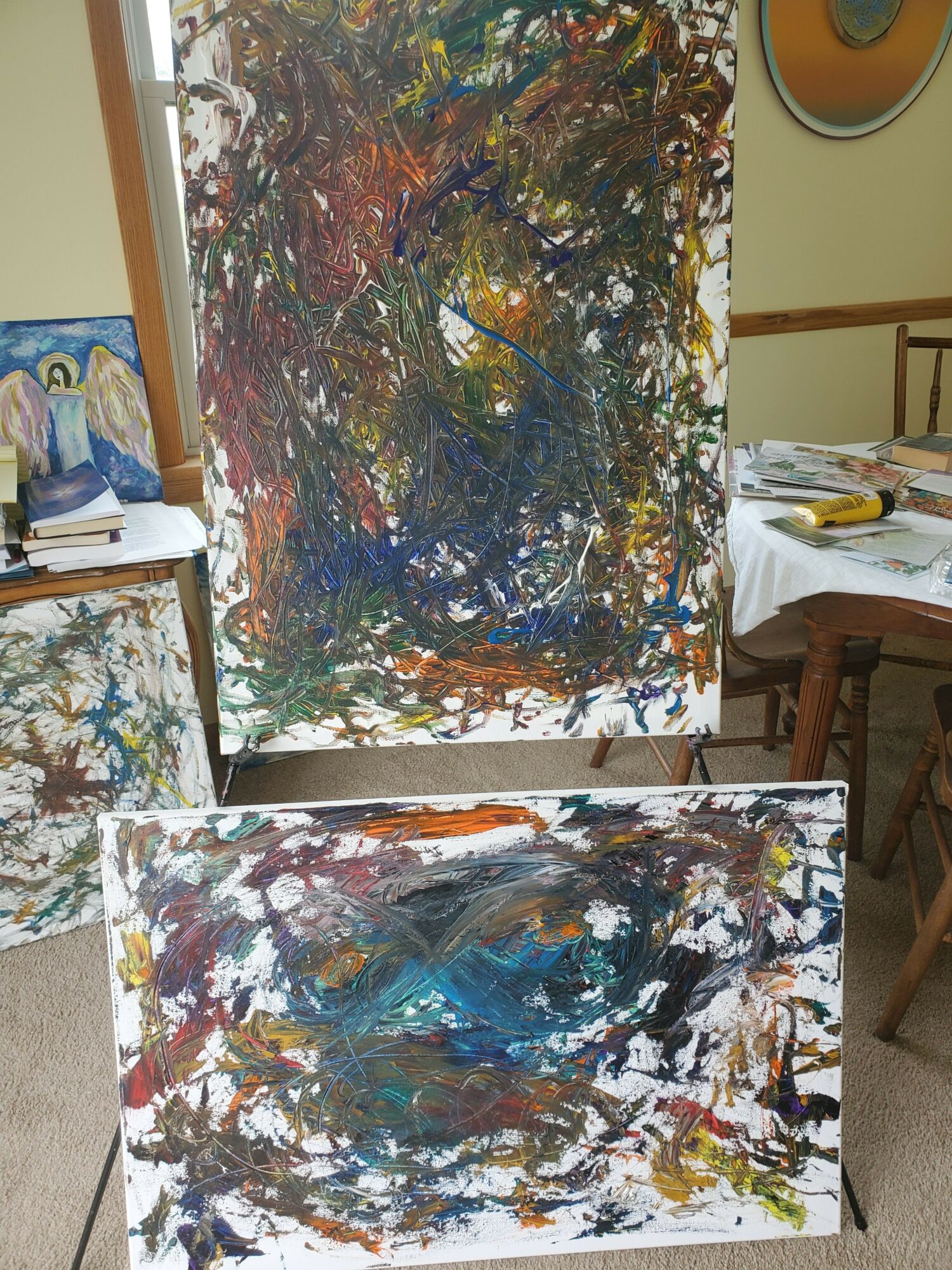Three abstract paintings on easels with colorful, textured brushstrokes in a room with wooden furniture.
