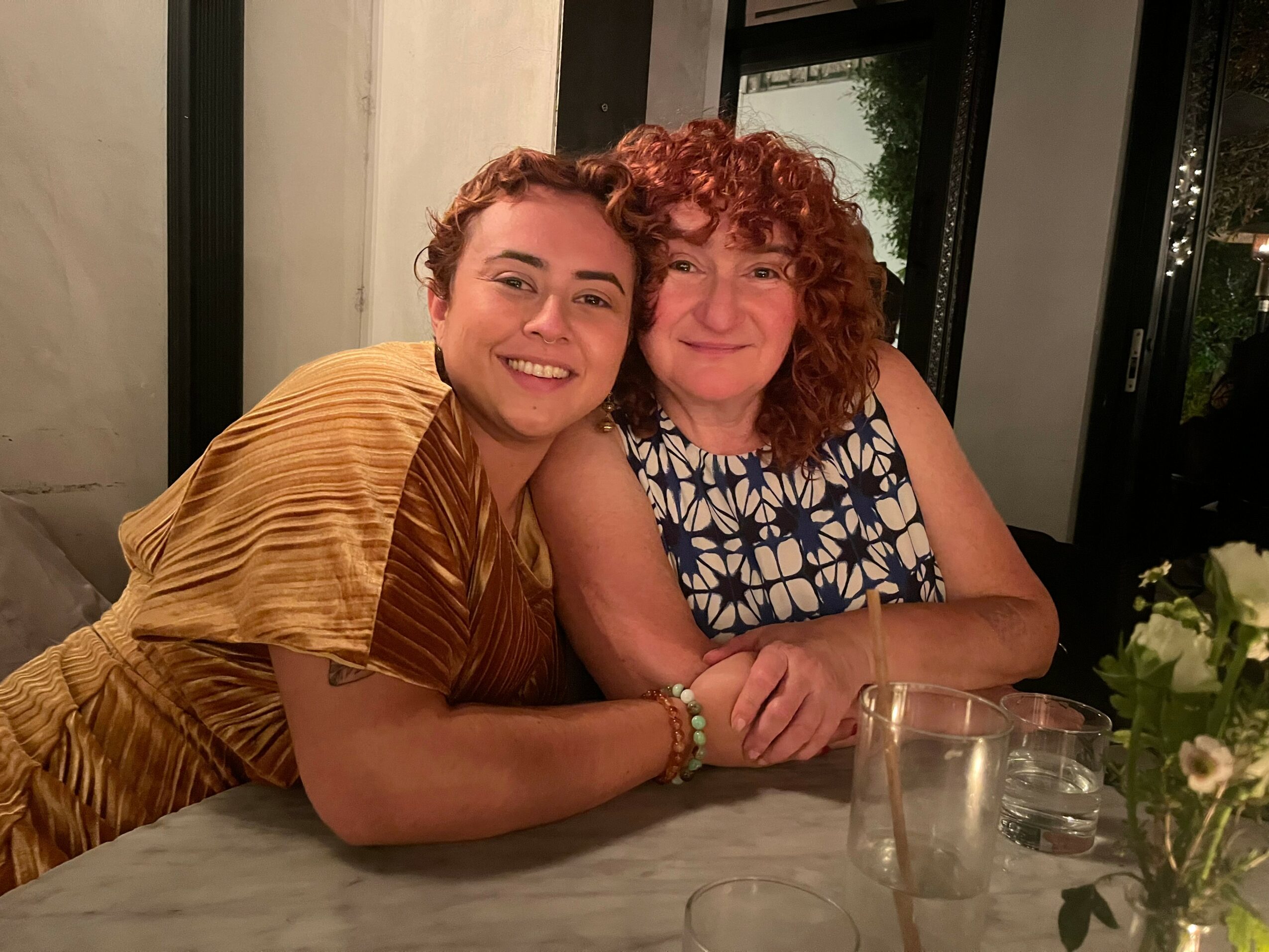 Two women smiling and sitting at a table with flowers and glasses, indoors with a window behind them.