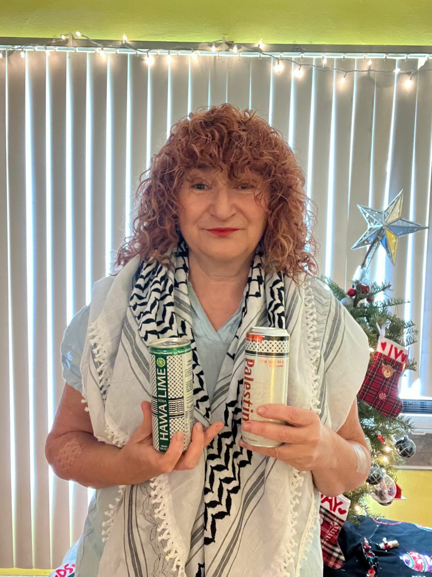 Woman with curly hair holding a drink, standing in front of a decorated Christmas tree, smiling.