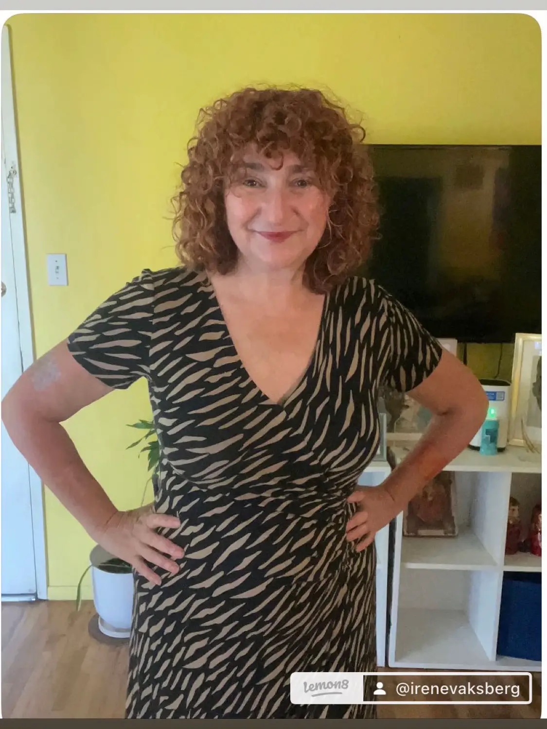 Woman with curly hair wearing a zebra-patterned dress, standing with hands on hips in a room with yellow wall and TV