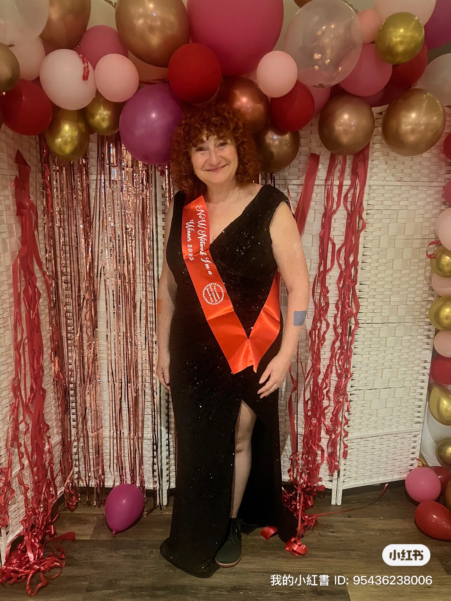 Woman with curly hair in a black dress stands in front of colorful balloons and decorations, smiling at the camera.