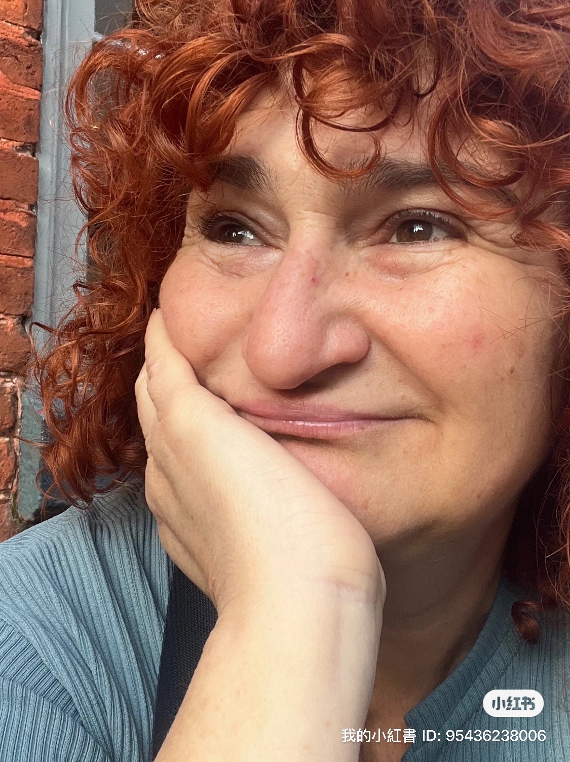 Close-up of a woman with curly red hair resting her face on her hand, smiling softly, outdoors.