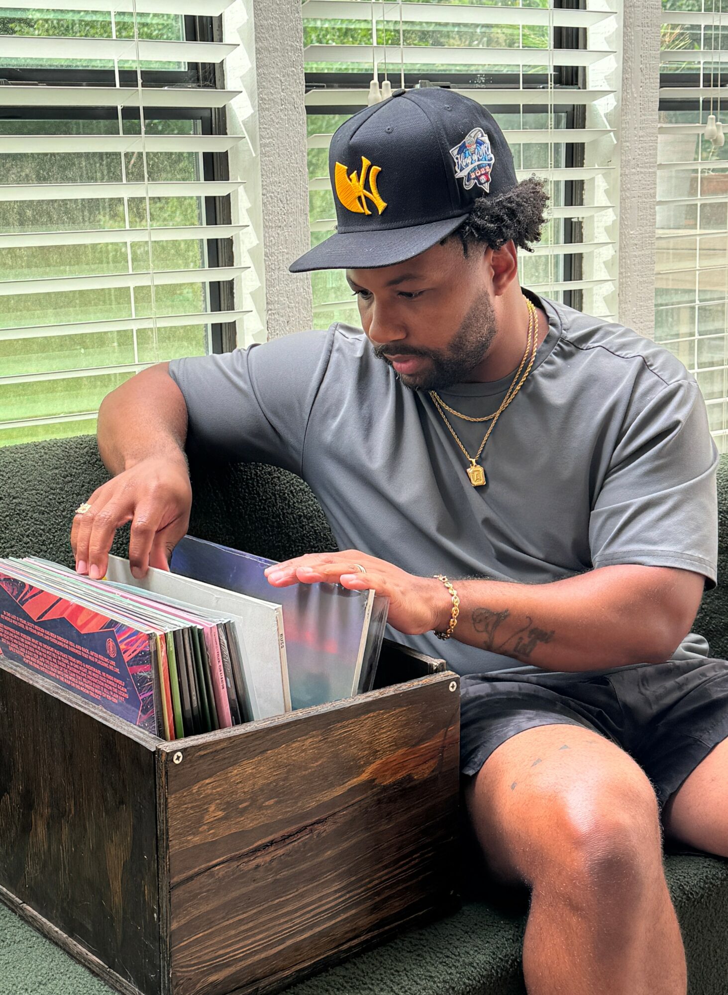 Young man with a cap, gray t-shirt, and shorts sitting by a window, looking through a box of records.