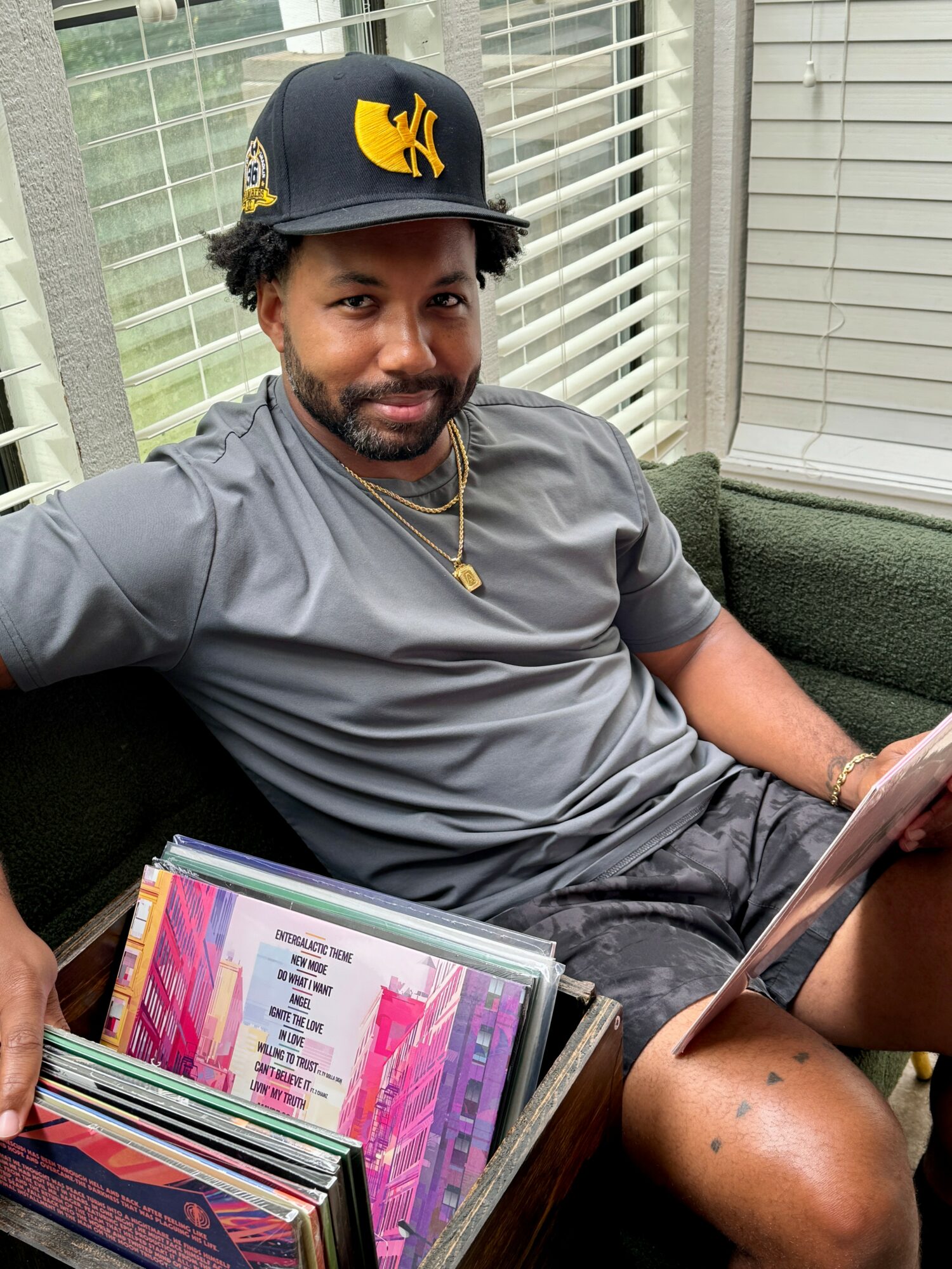 Man with a beard wearing a black cap with a yellow logo, gray shirt, and gold necklace, sitting on a green couch near window.