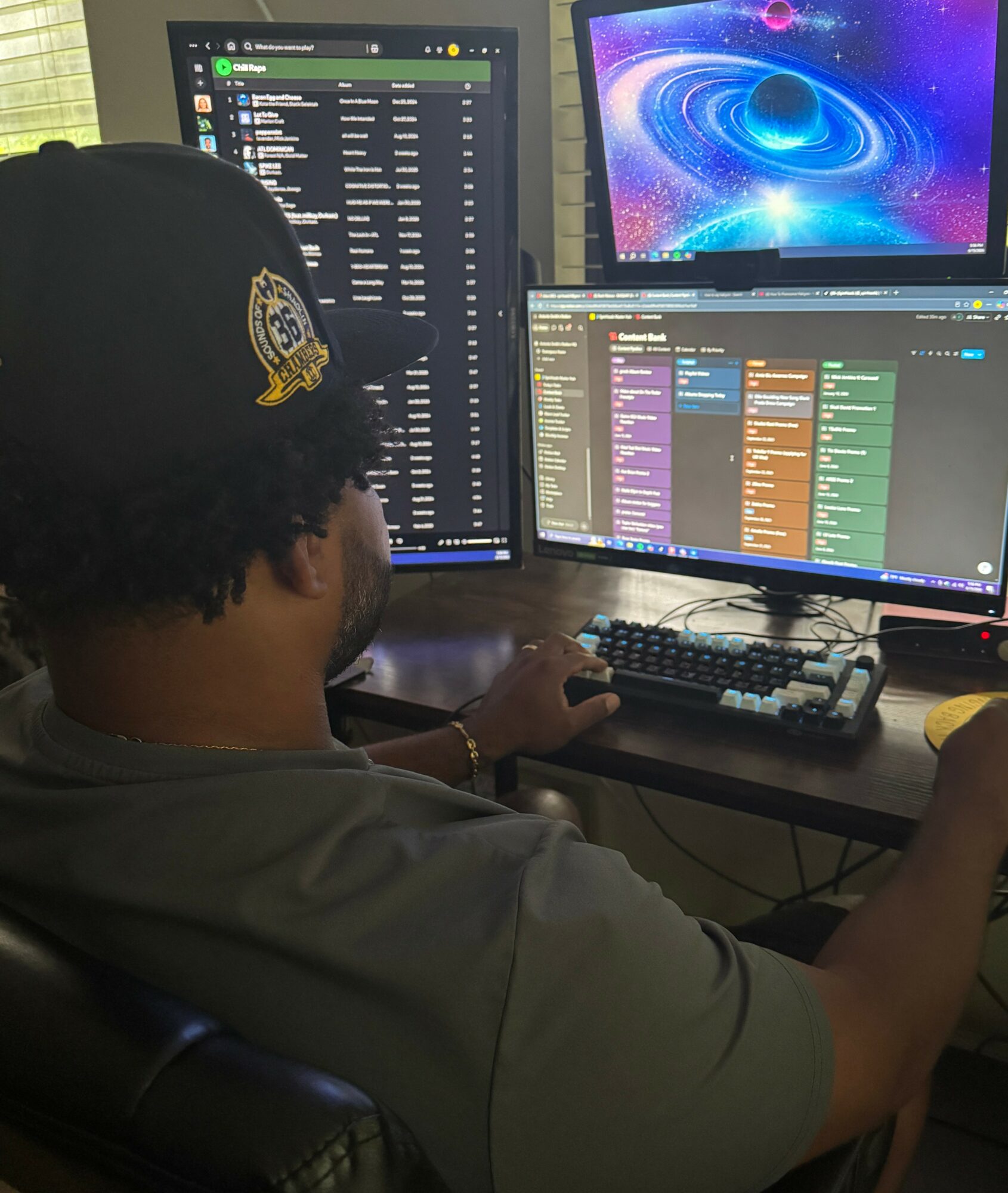 Person wearing a cap working at a desk with three monitors displaying music or media applications.