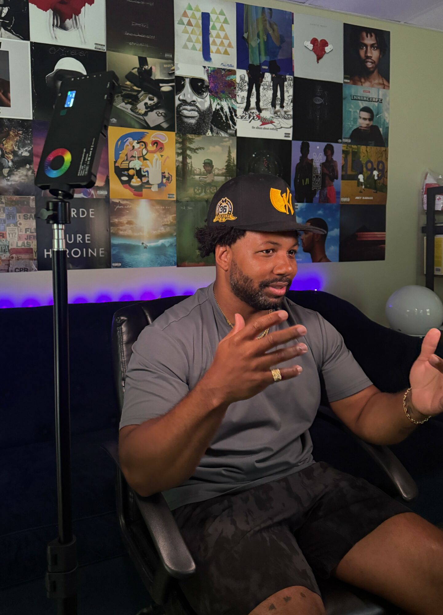 Man sitting in a chair, gesturing with hands, wearing a cap, in a room with a wall of posters and photos