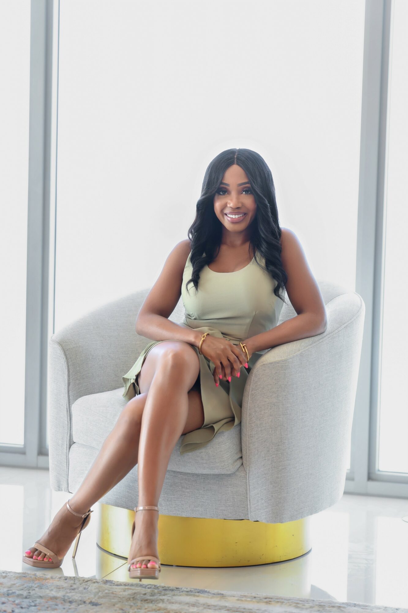 Woman sitting on a light-colored armchair in front of large windows, smiling, with crossed legs and wearing a sleeveless dress.