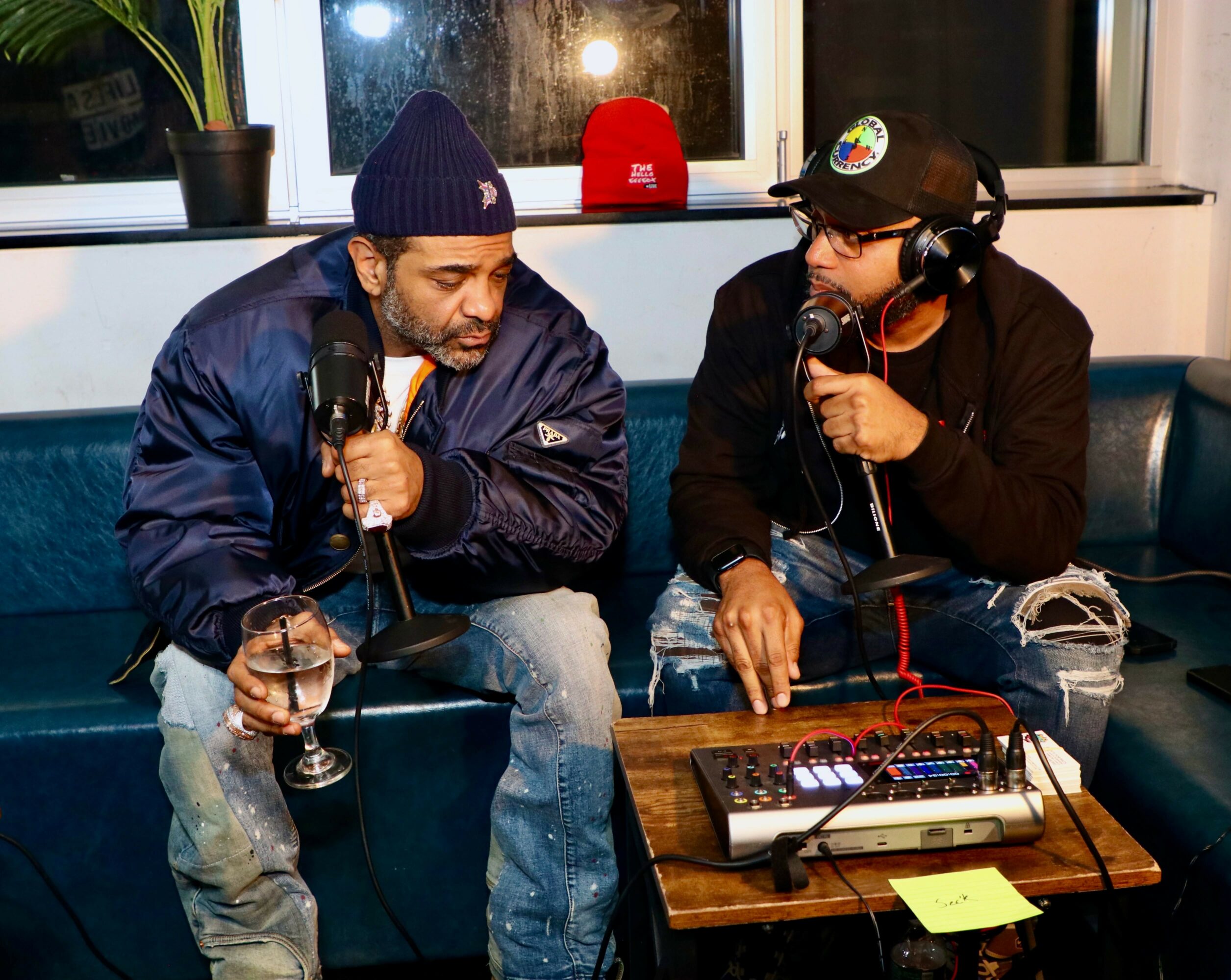 Two men sit on a couch with microphones, one holding a glass, a sound mixer on a small table, behind a window.