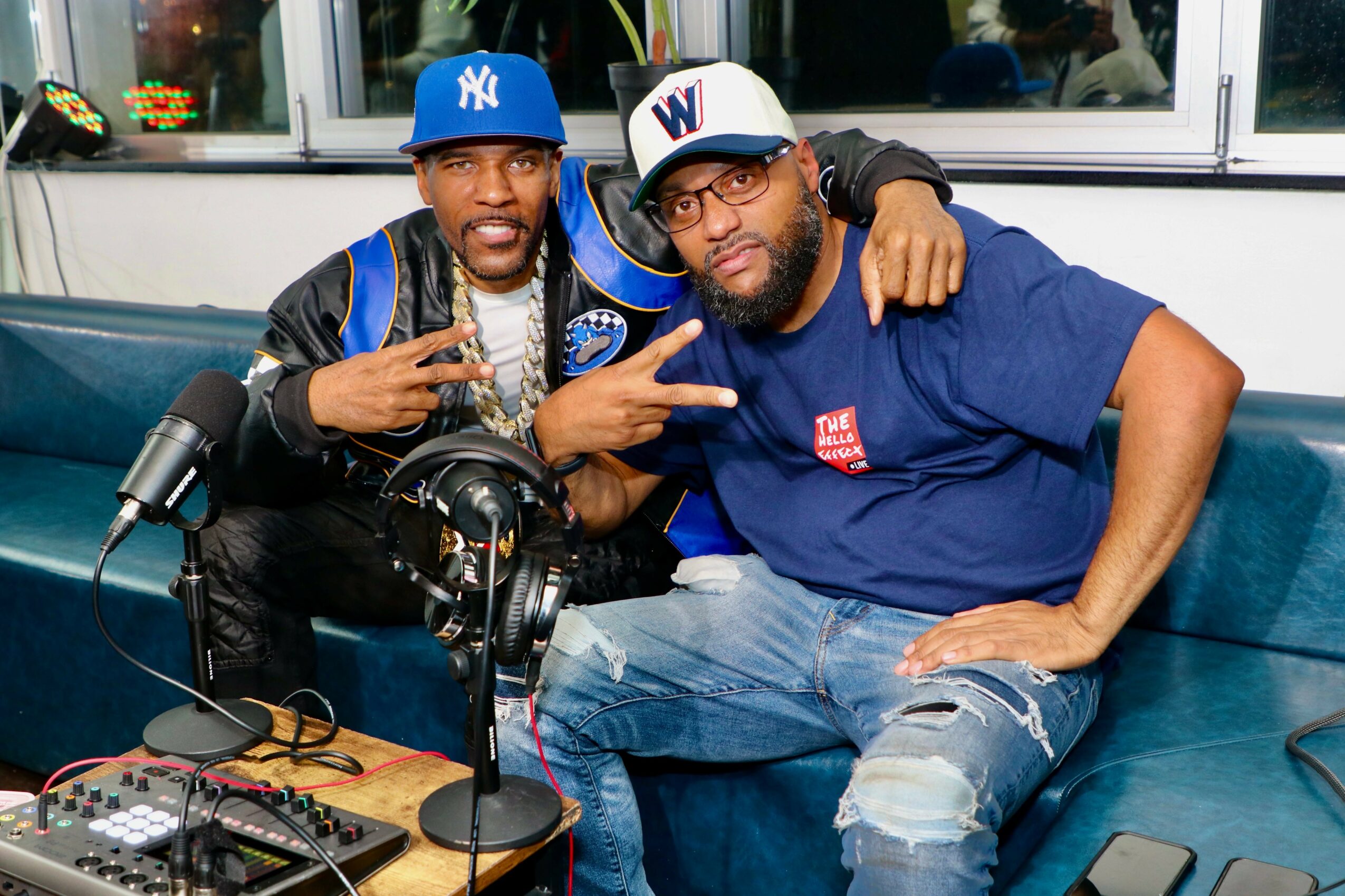 Two men sitting on a blue couch, one wearing a blue cap and the other a white cap, with a microphone and audio equipment in front of them.