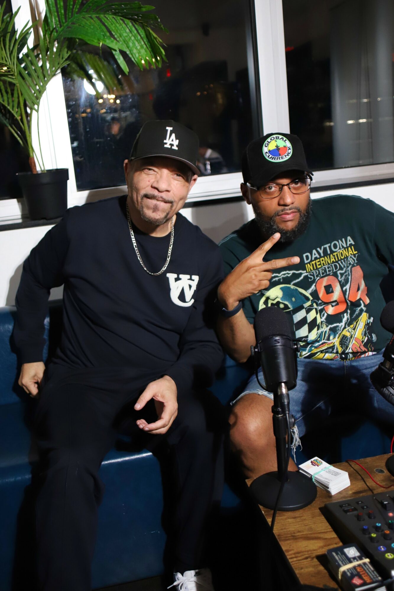 Two men sitting indoors, one wearing a black cap and black sweatshirt, the other wearing glasses and a colorful T-shirt, with a microphone in front.