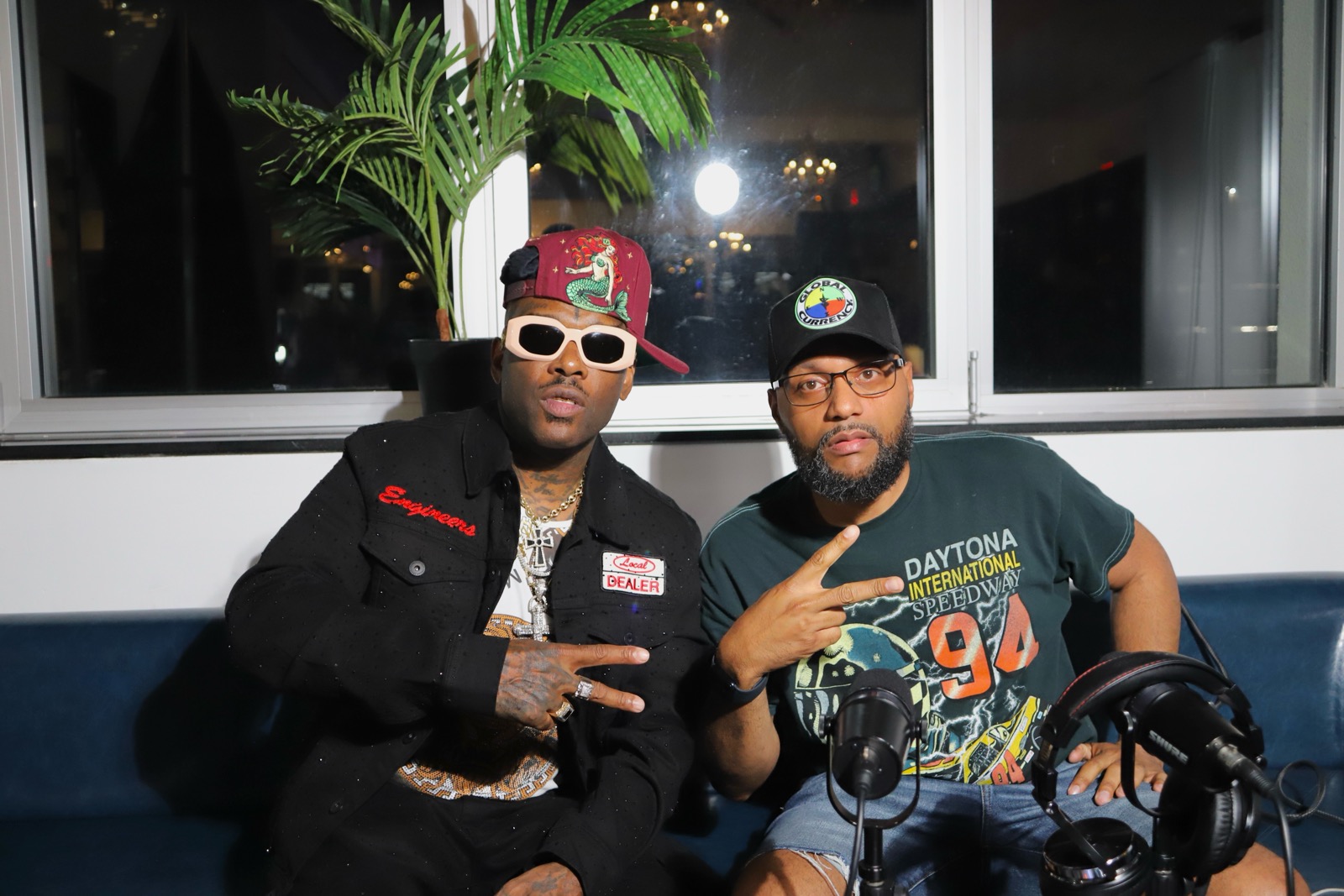 Two men sitting indoors in front of a window, one wearing a colorful cap and sunglasses, the other wearing a cap and glasses, both making peace signs.