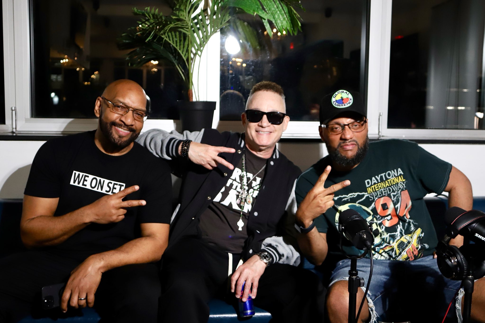 Three men sitting together indoors, smiling, making peace signs, with a large plant and window behind them.