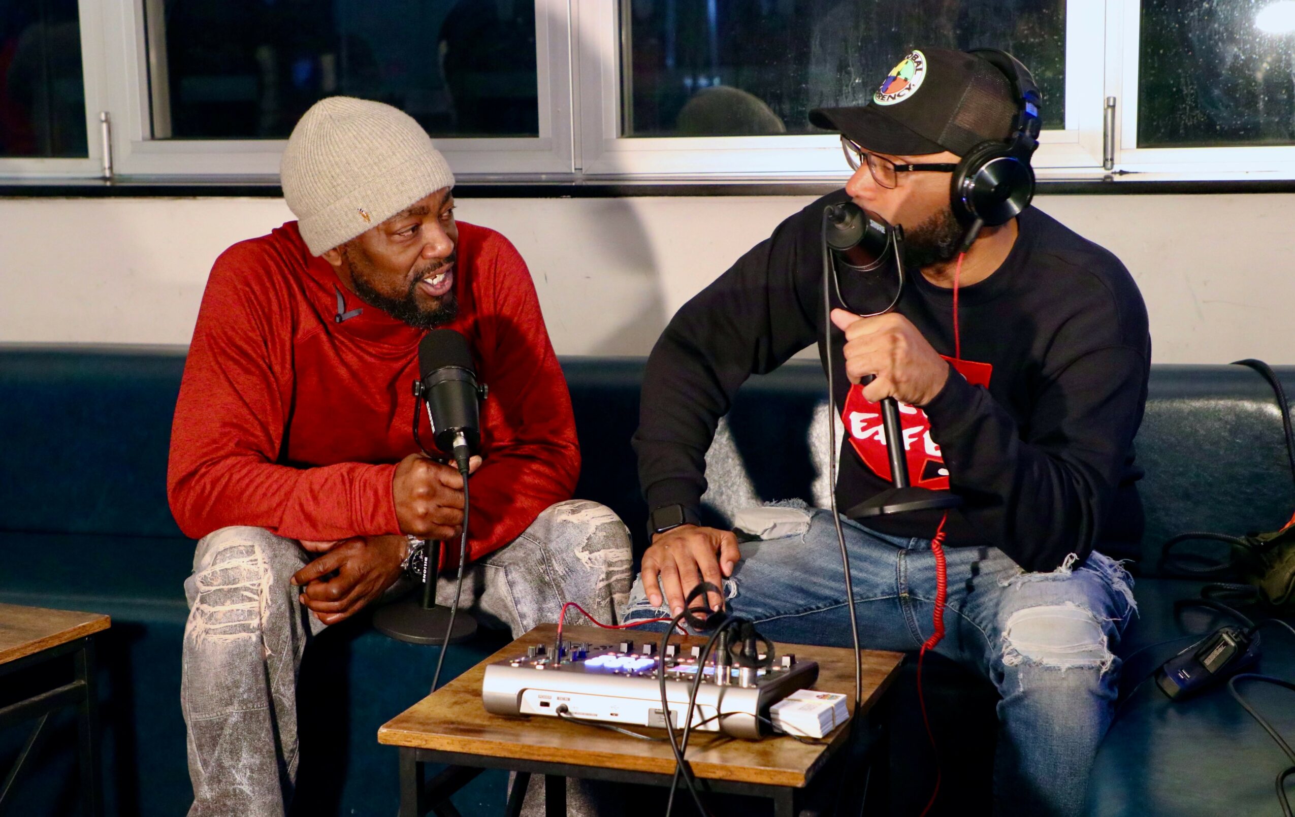 Two men sit on a couch with microphones, recording a podcast or radio show, with audio equipment on a table between them.